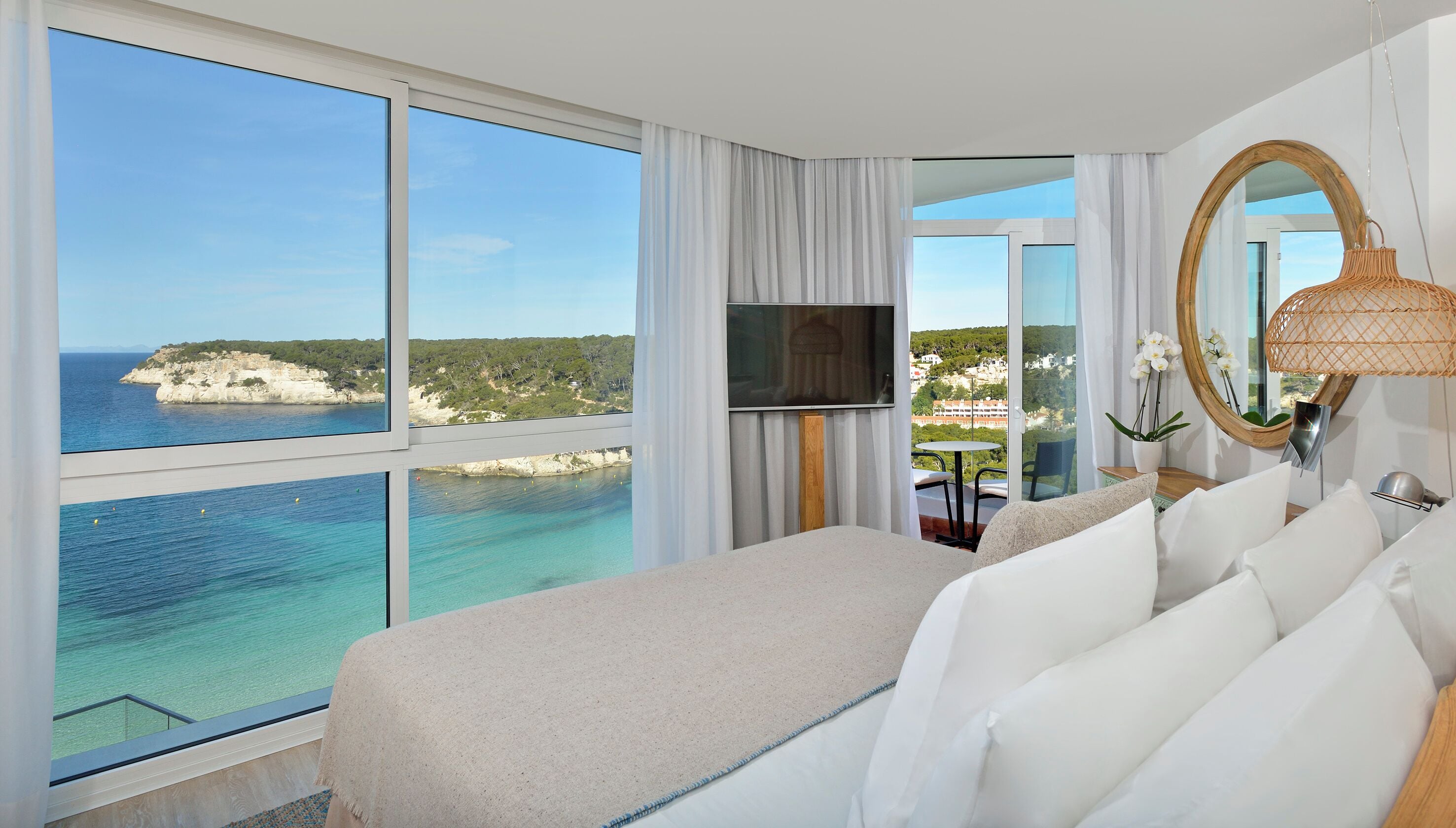 Habitación de hotel con un ventanal grande que ofrece vista al mar, cama con ropa blanca, televisor y espejo redondo.