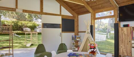 A spacious room with a high ceiling, wooden beams, a round table with chairs, a bookshelf, a play area with a tent and toys, and a view of a garden through large windows.