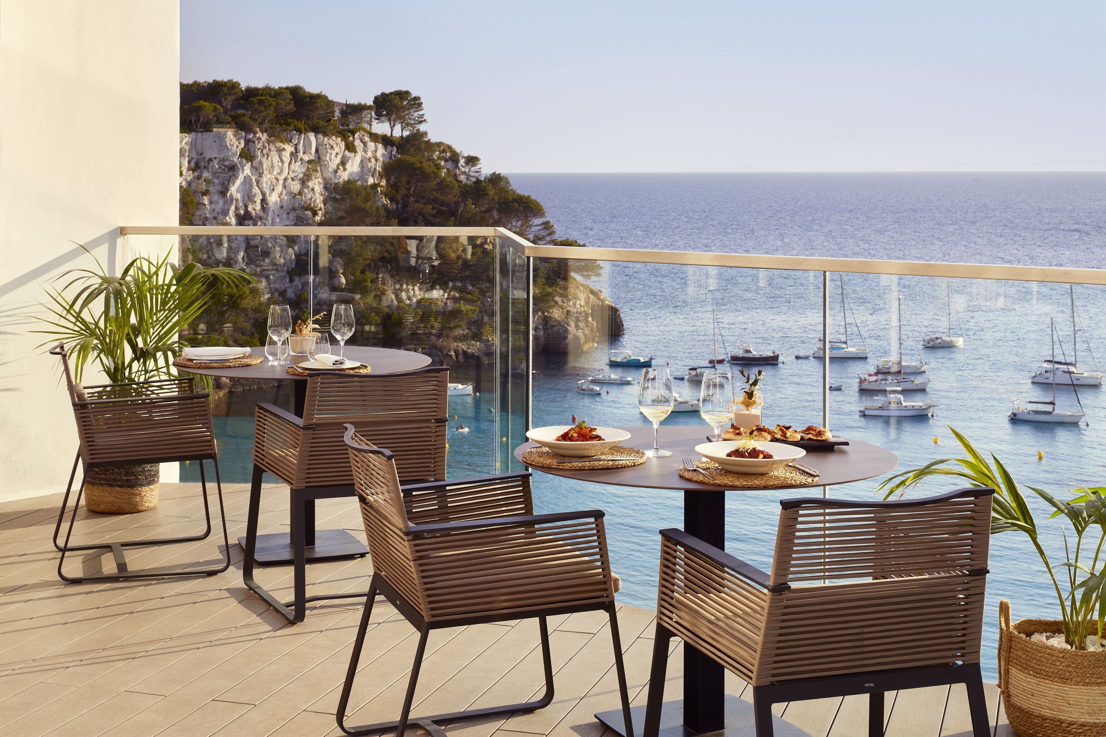 A terrace with a table set for dining, overlooking a coastal area with boats and cliffs.