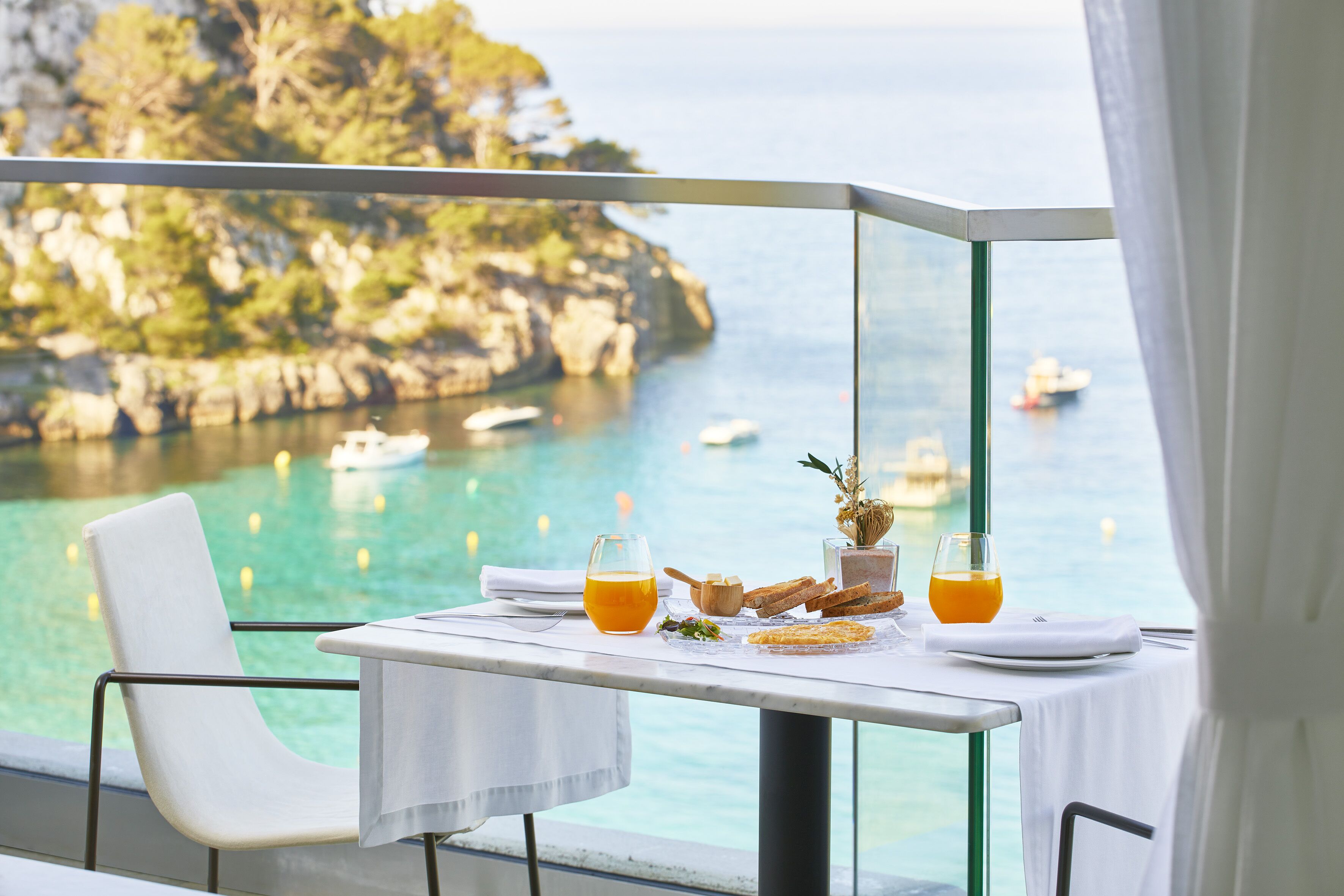 A table with breakfast items and drinks, overlooking a coastal view with boats and cliffs.