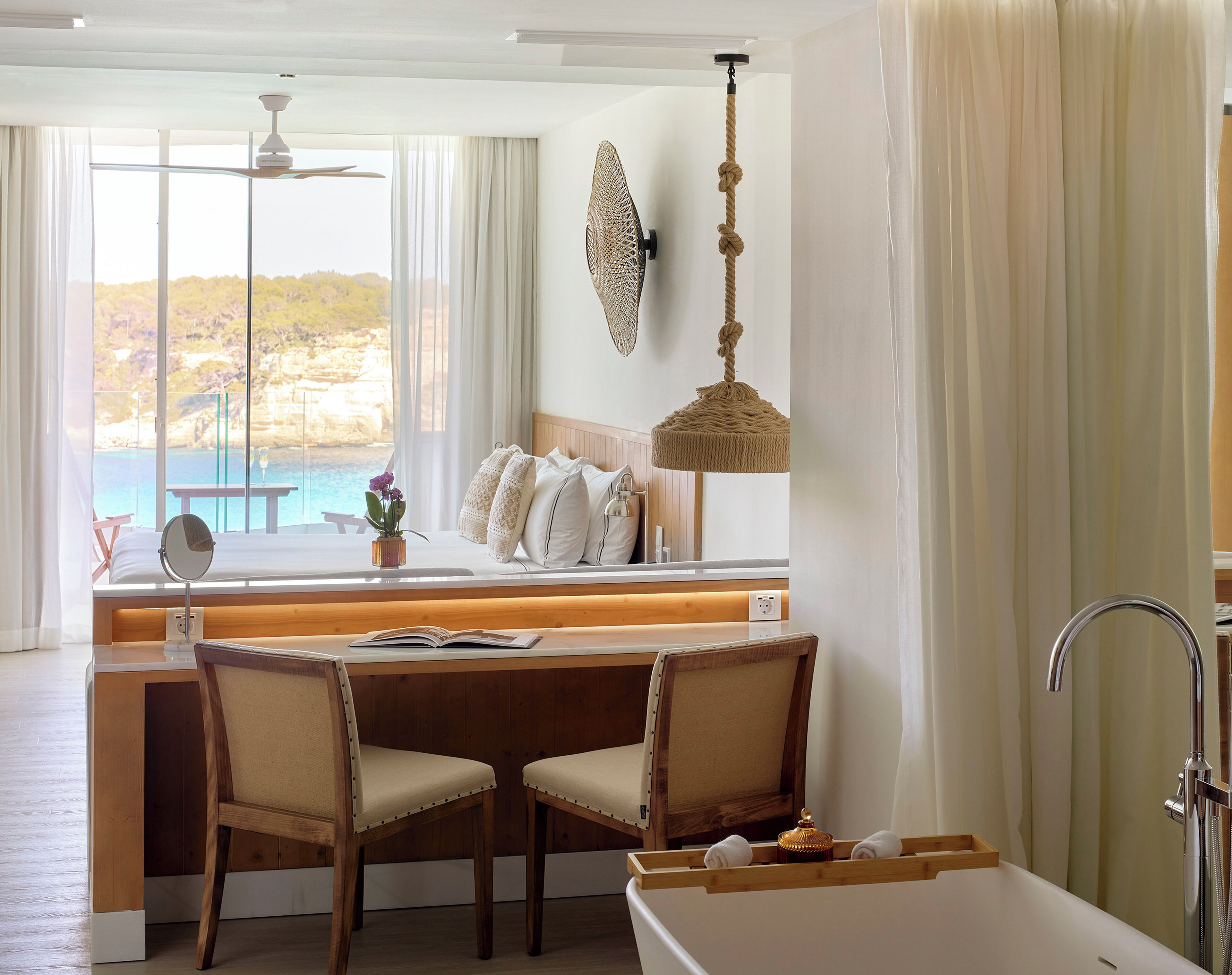 Habitación de hotel con un gran ventanal, un escritorio de madera con dos sillas, cama con ropa blanca y una bañera.