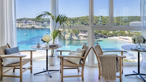 Habitación con muebles de mimbre, una planta en maceta y vistas a la costa con barcos y una playa.