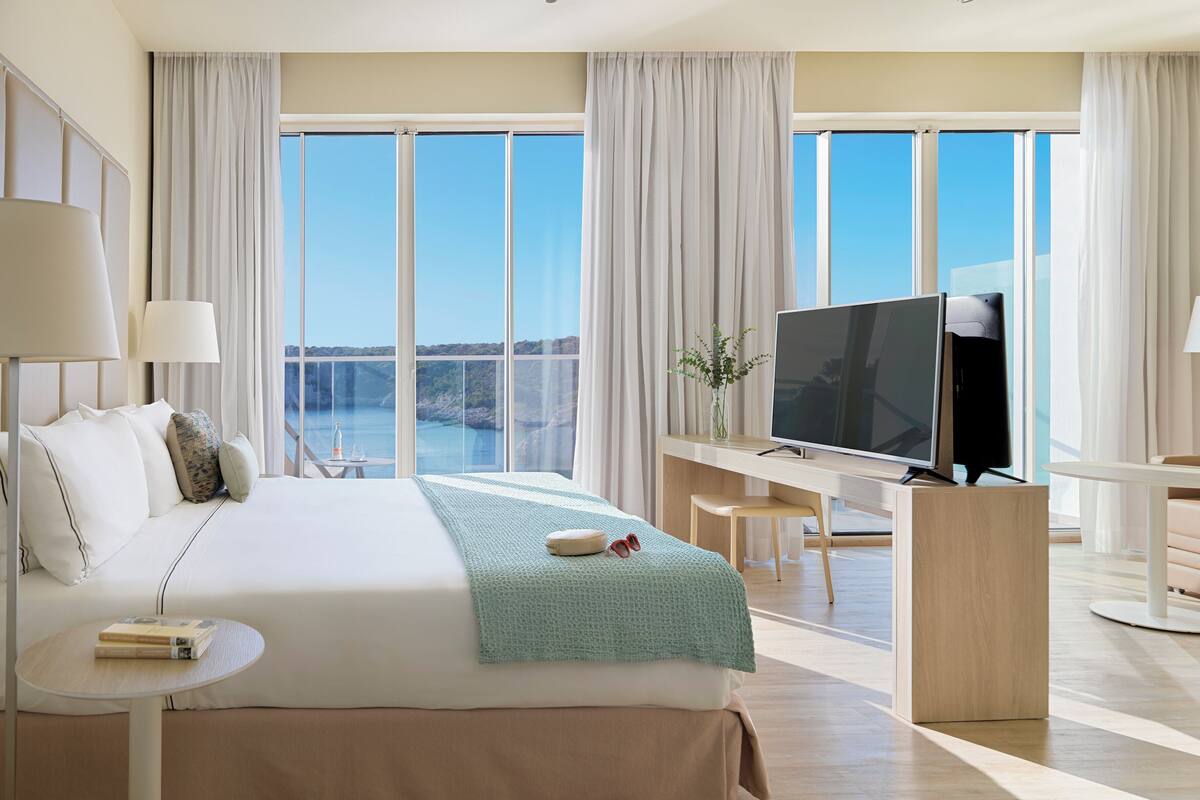 Ein modernes Hotelzimmer mit einem großen Bett, einem Schreibtisch mit Fernseher, Meerblick und einem Balkon.