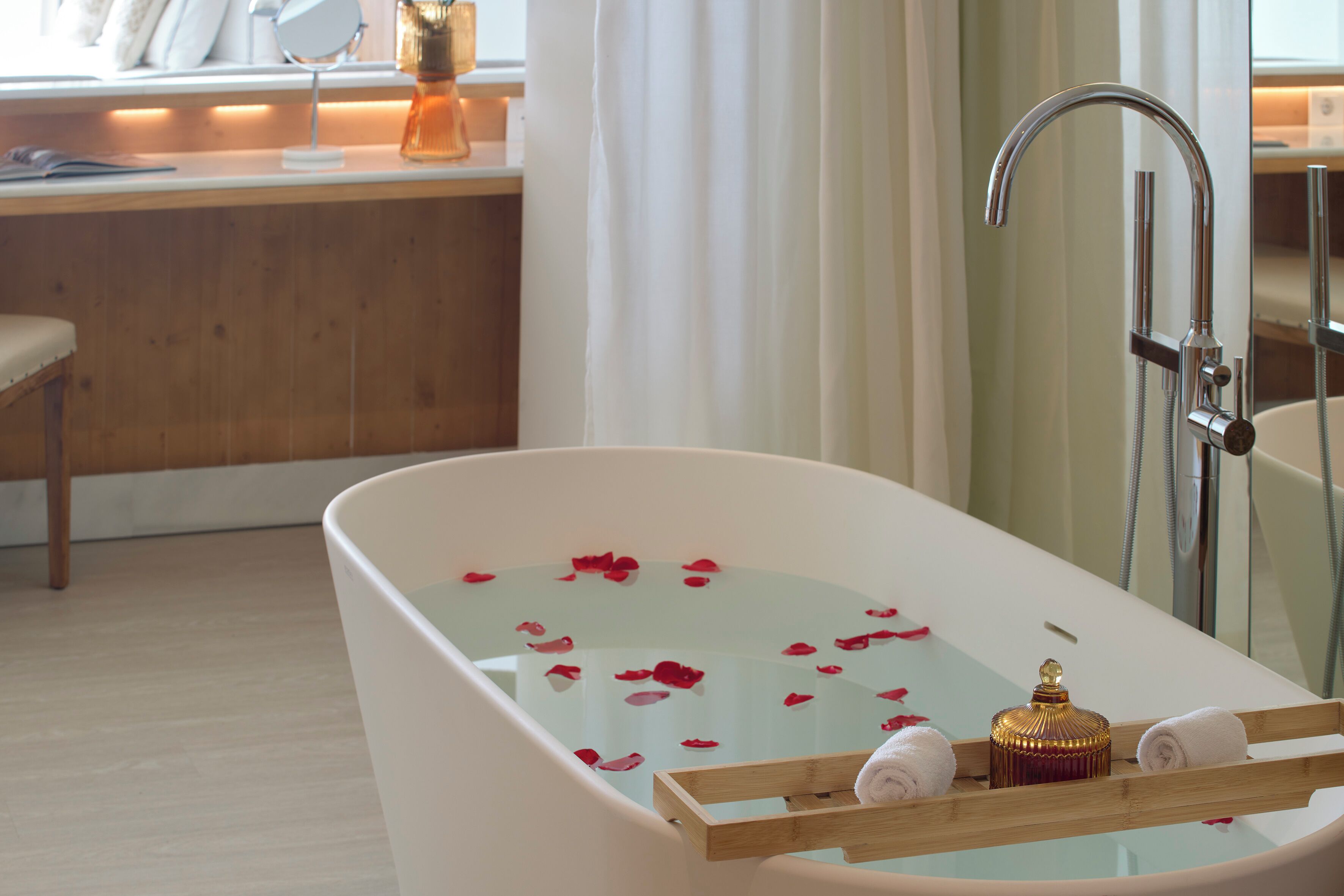 A modern bathroom with a freestanding bathtub, a wooden tray with towels, and rose petals in the water.