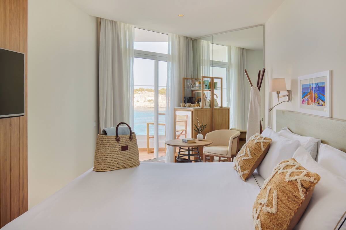 Ein Hotelzimmer mit Bett, Fernseher, Schreibtisch mit Stuhl, einem kleinen Tisch und Meerblick durch große Fenster.