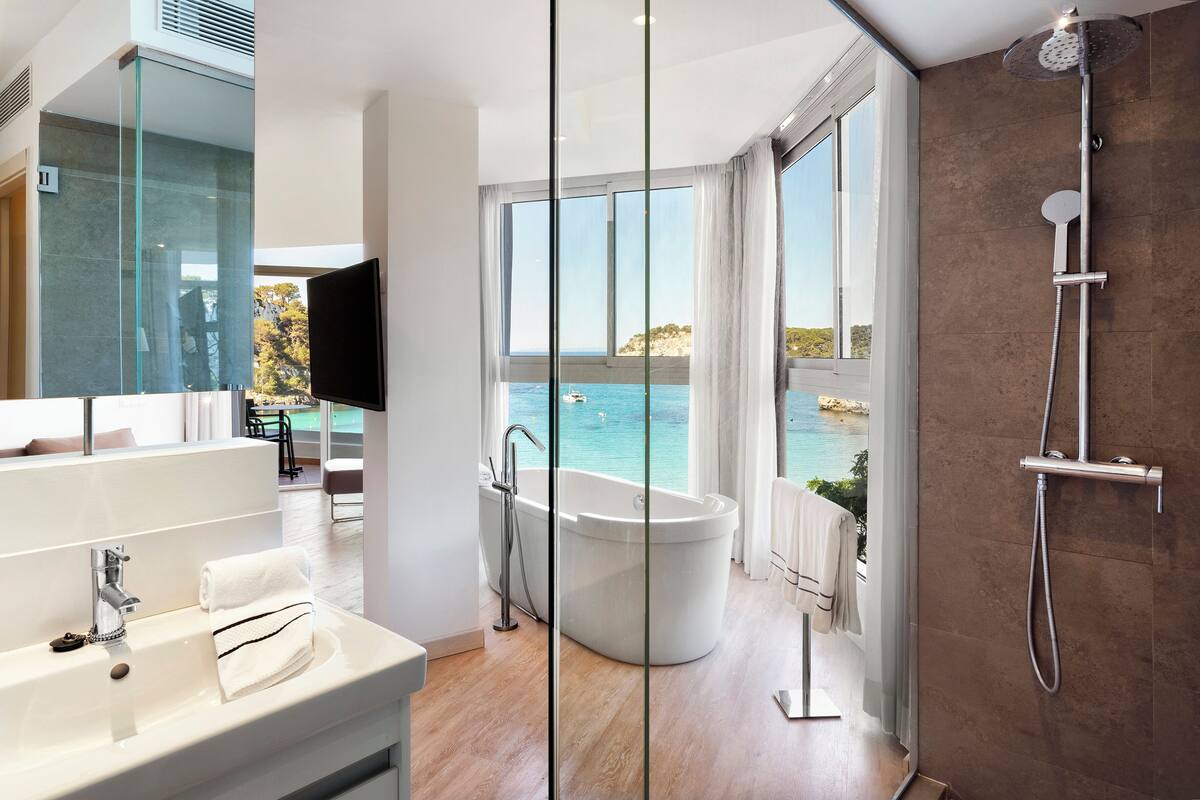 Ein modernes Badezimmer mit einer großen Badewanne, einer separaten Duschkabine und einem großen Fenster mit Meerblick.
