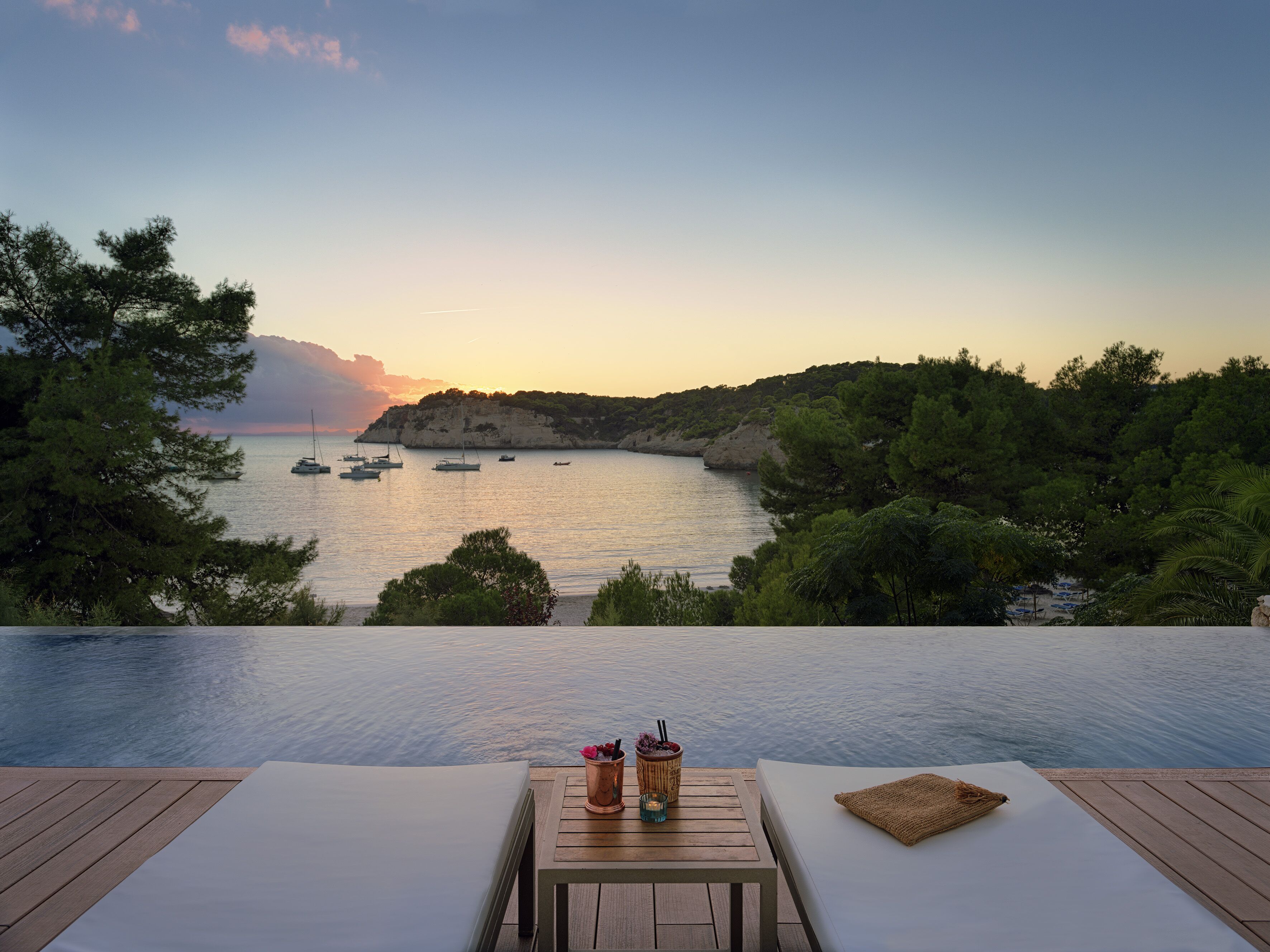 Une piscine à débordement offrant une vue sur les bateaux sur l’eau, une table avec des boissons et des chaises longues.