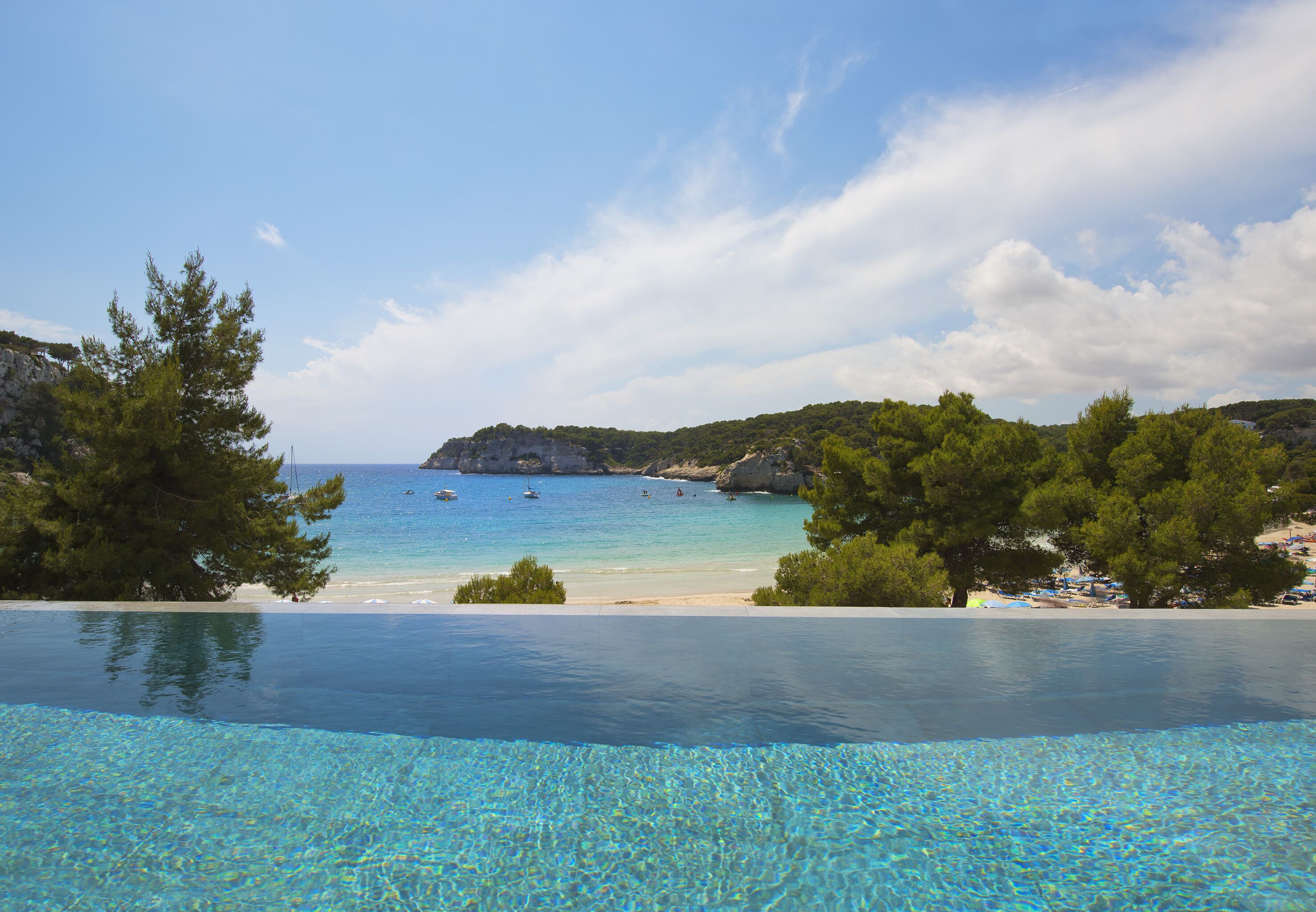 Une piscine à débordement à l’eau d’un bleu cristallin, une plage et un paysage montagneux en toile de fond.