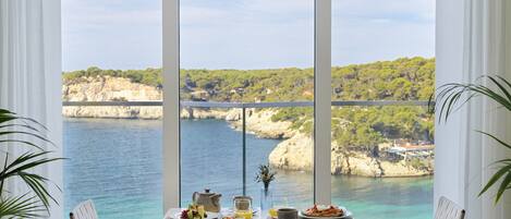 A dining area with a table set for two, overlooking a coastal landscape with cliffs and greenery.
