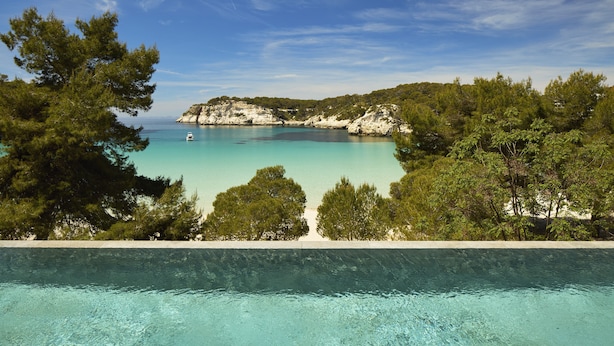 Una piscina con agua turquesa cristalina, rodeada de árboles y vistas a un paisaje costero con acantilados y un barco a lo lejos.