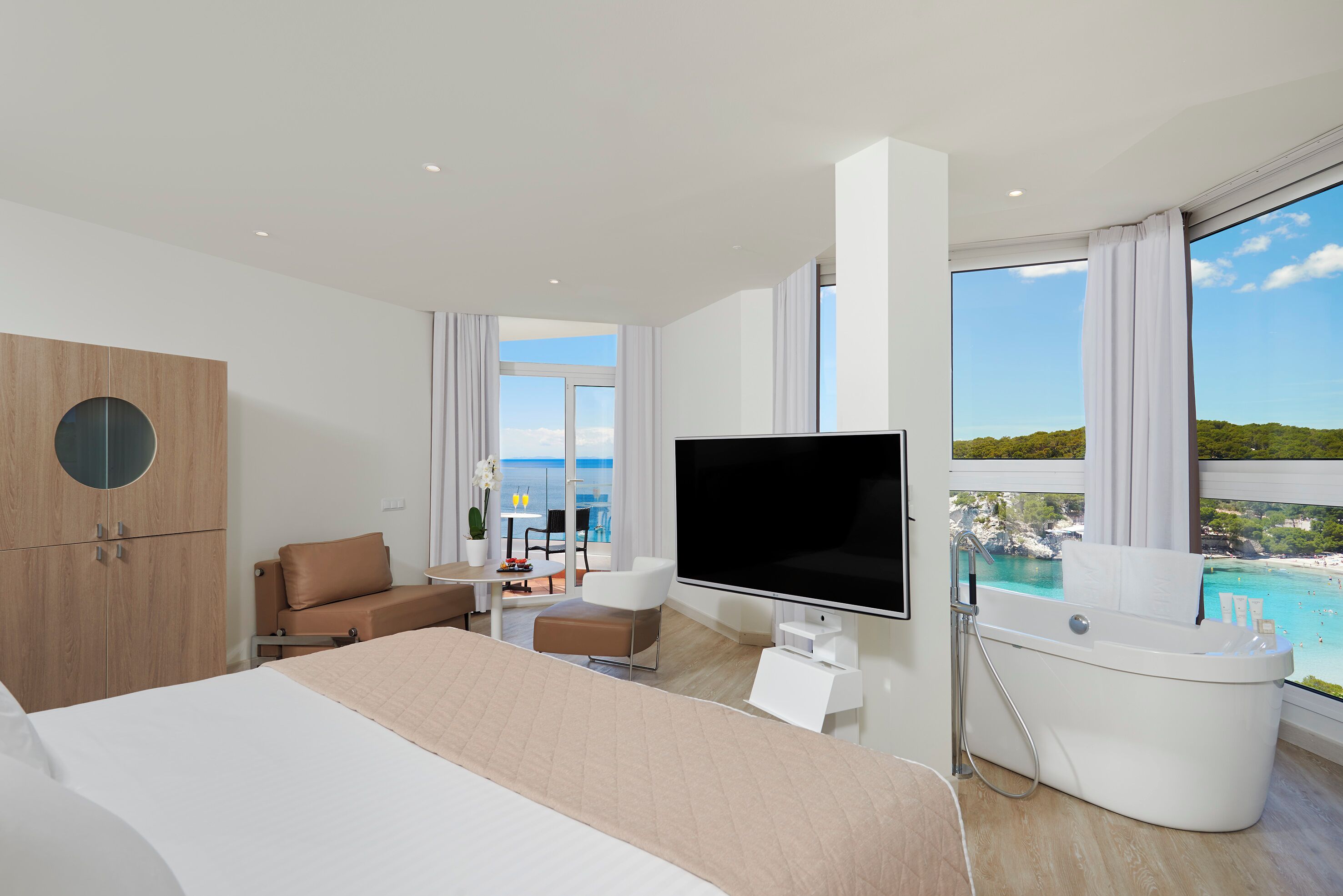 Habitación de hotel moderna con una cama grande, un televisor de pantalla plana, una bañera y un balcón con vista al mar.