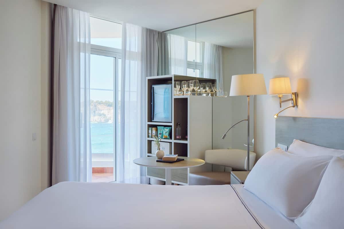 Ein Hotelzimmer mit Bett, Schreibtisch mit Stuhl, Bücherregal und einem großen Fenster mit Vorhängen.
