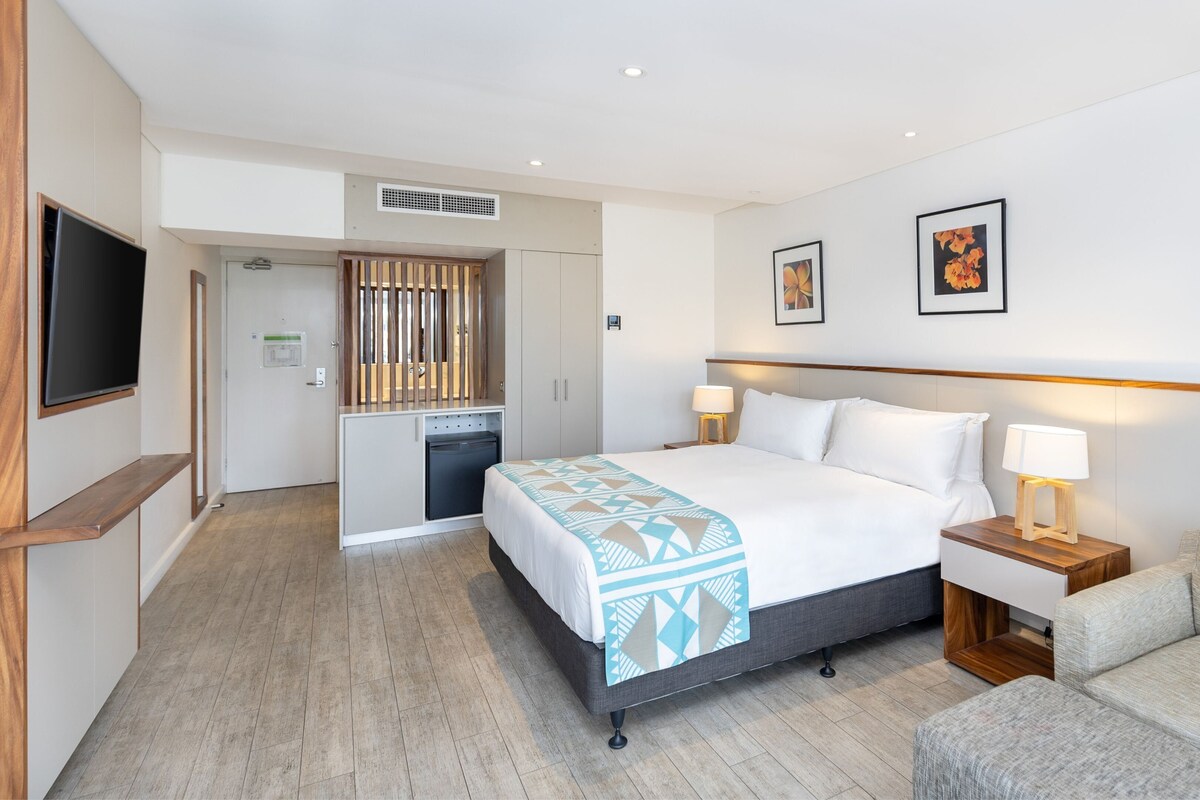 Una habitación de hotel con cama, mesitas de noche, televisor, escritorio y una silla.