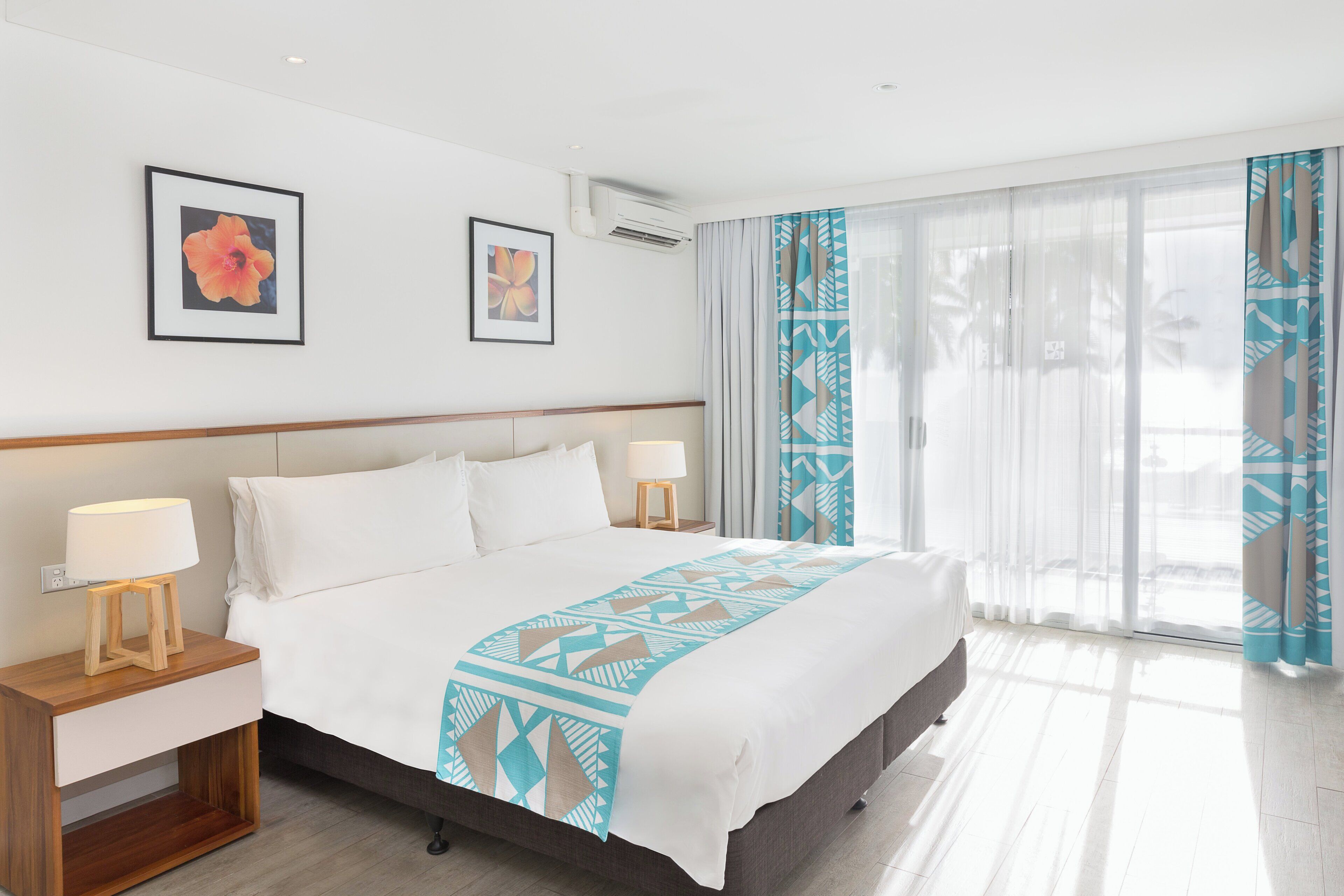 A hotel room with a large bed, two bedside tables with lamps, a window with sheer curtains, and two framed floral artworks on the wall.