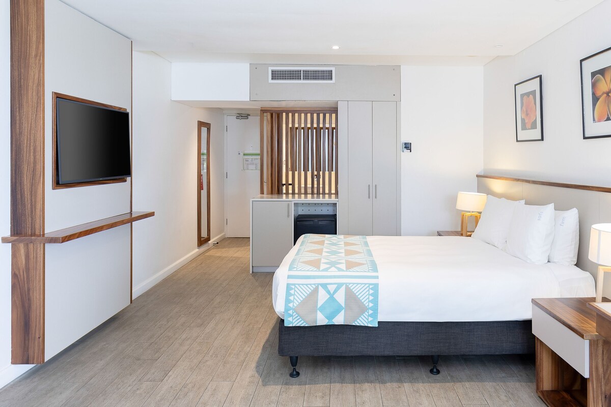 Habitación de hotel con cama, mesitas de noche, televisor, escritorio y kitchenette.