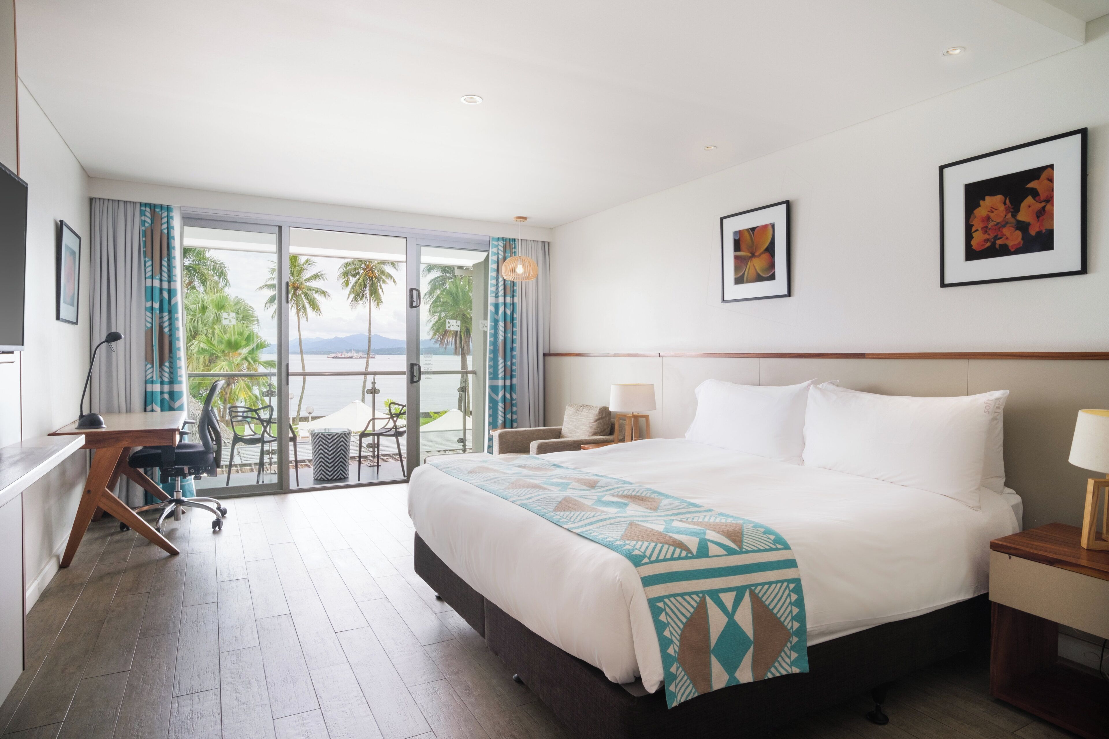 A hotel room with a bed, a desk, a chair, a nightstand, and a view of palm trees and water.