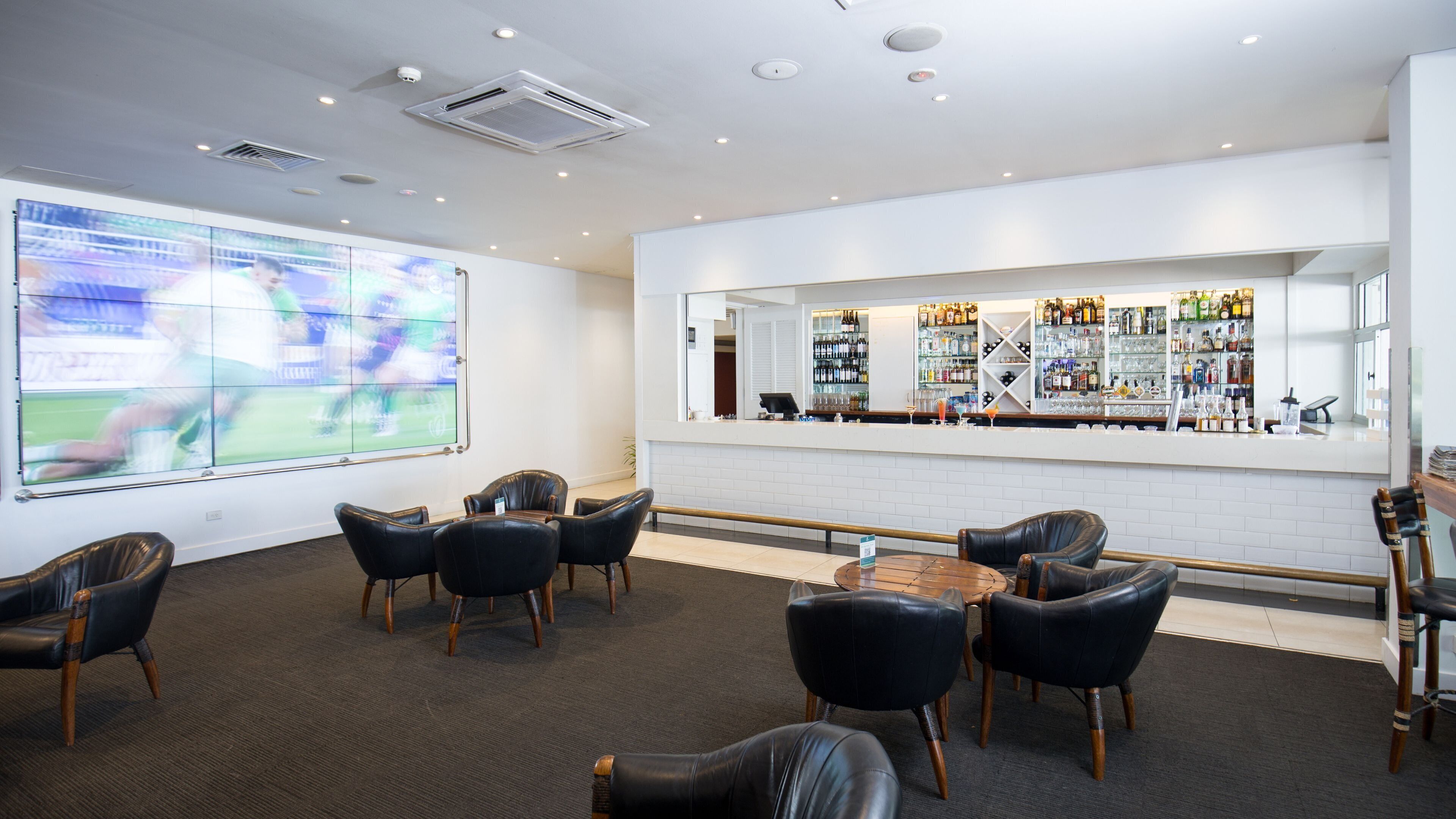 A bar area with leather chairs, a counter with bottles, and a large TV screen displaying a sports event.