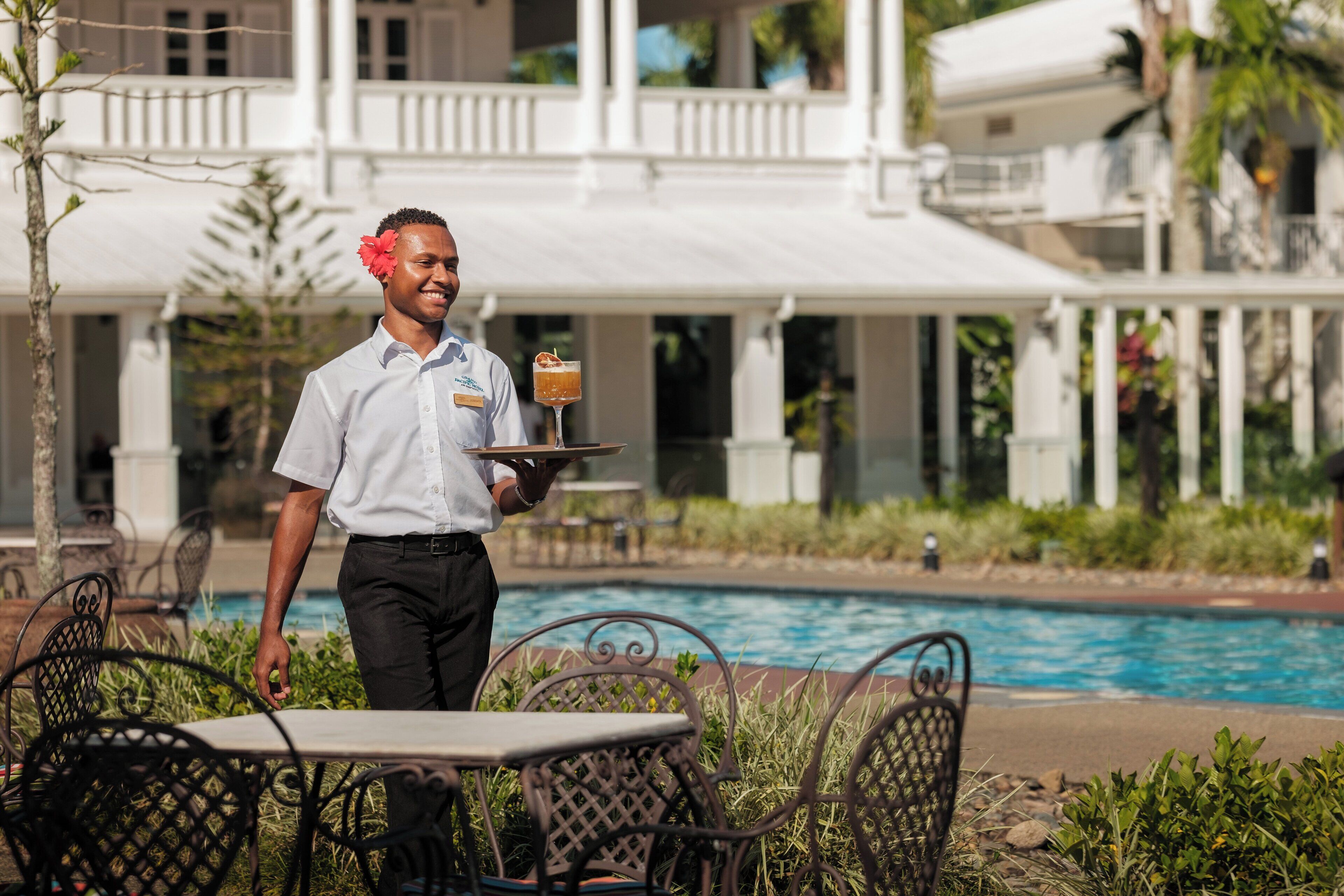 A waiter carrying a tray with a drink and a dessert, standing near a pool with outdoor seating.