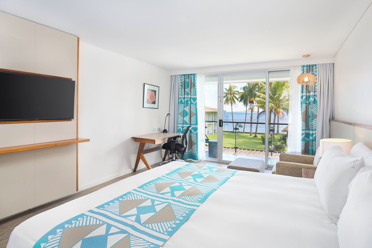 Habitación de hotel con cama, escritorio, silla, televisión y balcón con vistas a palmeras y al océano.