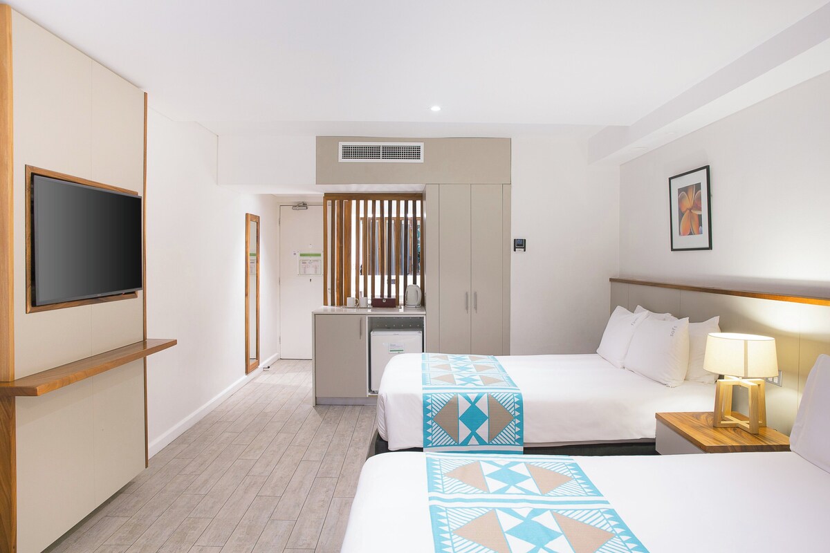 Habitación de hotel con dos camas, televisión, escritorio con computadora y kitchenette.