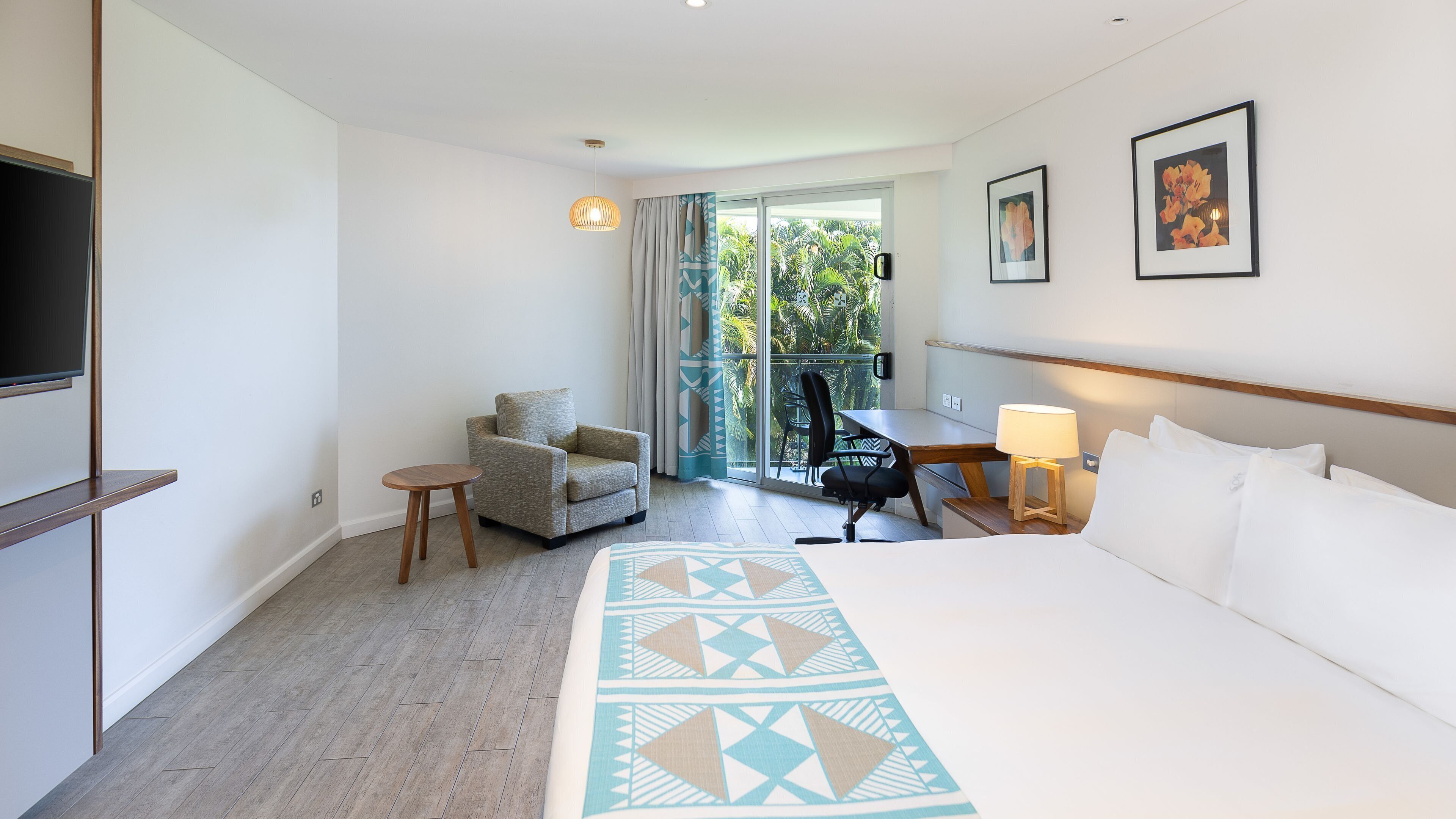 A hotel room with a bed, a chair, a desk, a TV, and a balcony with a view of palm trees.