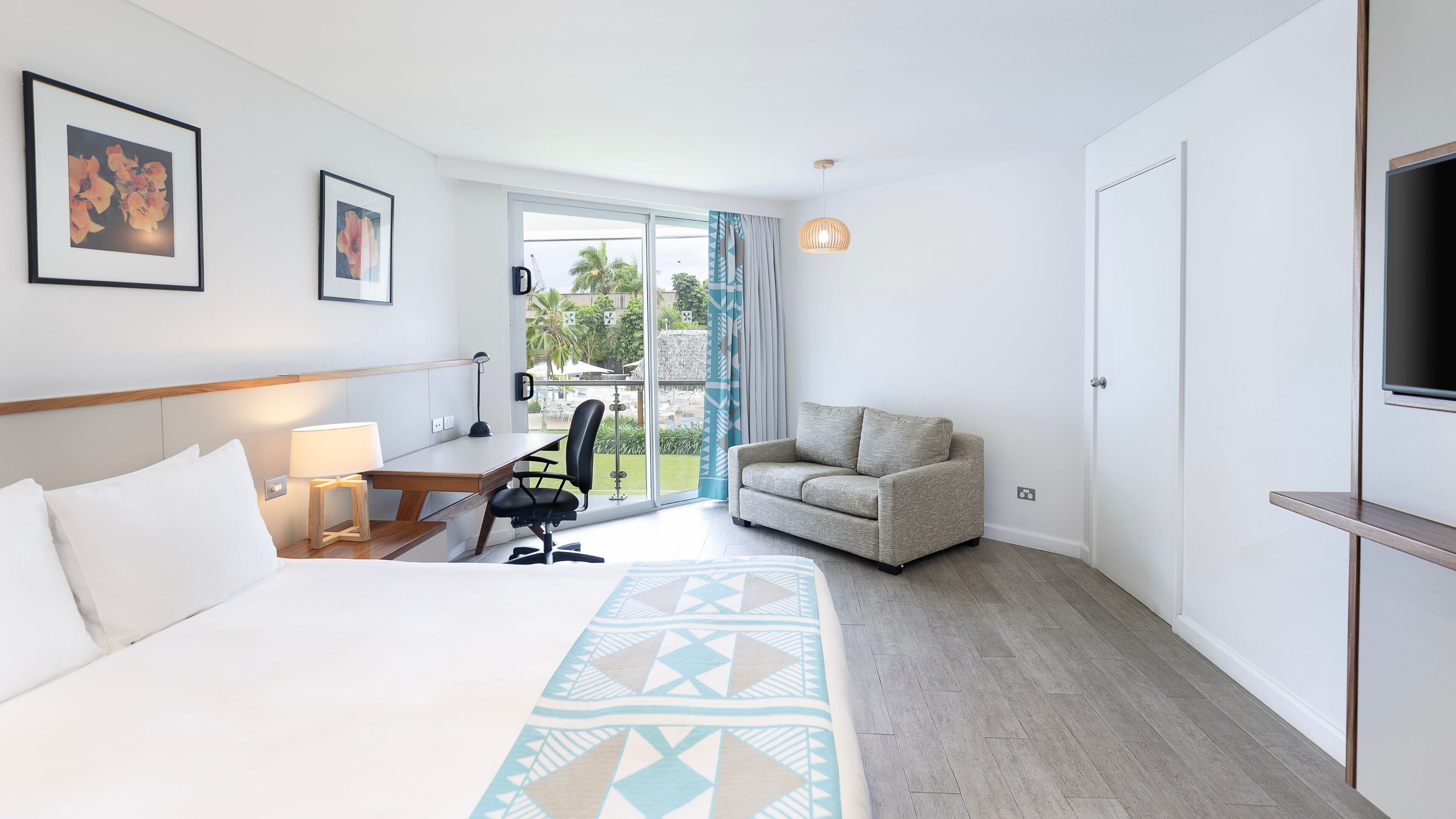 Hotelkamer met een bed, een bureau met stoel, een bank, een televisie en een balkon met uitzicht op groen.