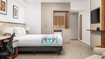 A hotel room with a bed, desk, chair, and a small kitchenette area.