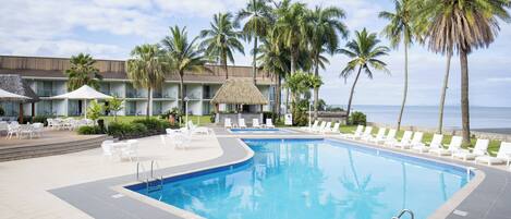 A swimming pool with lounge chairs, palm trees, and a building with a thatched roof.