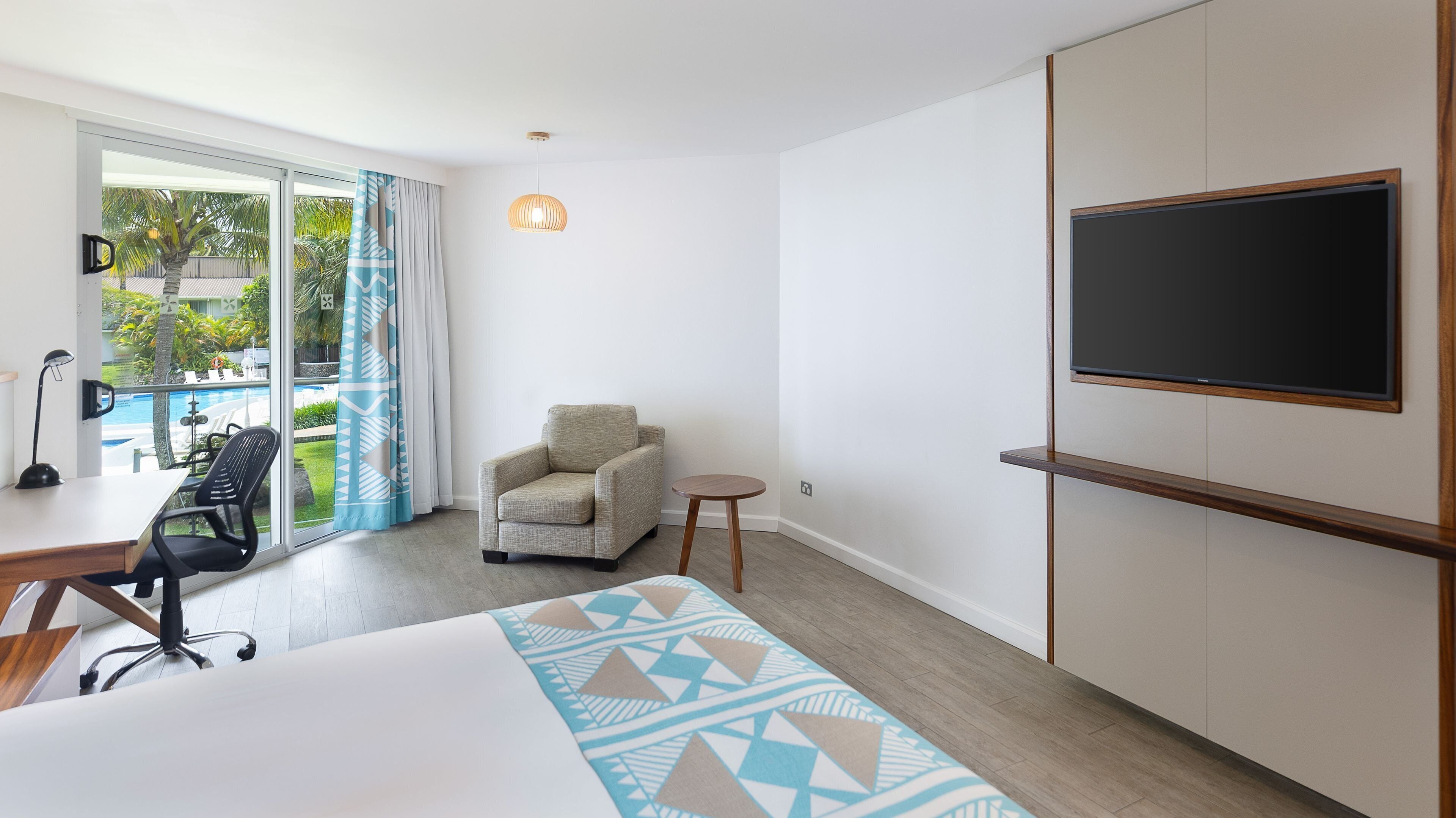 A hotel room with a bed, a desk with a chair, a small round table, a sofa, a TV mounted on the wall, and a view of a pool and greenery through the window.