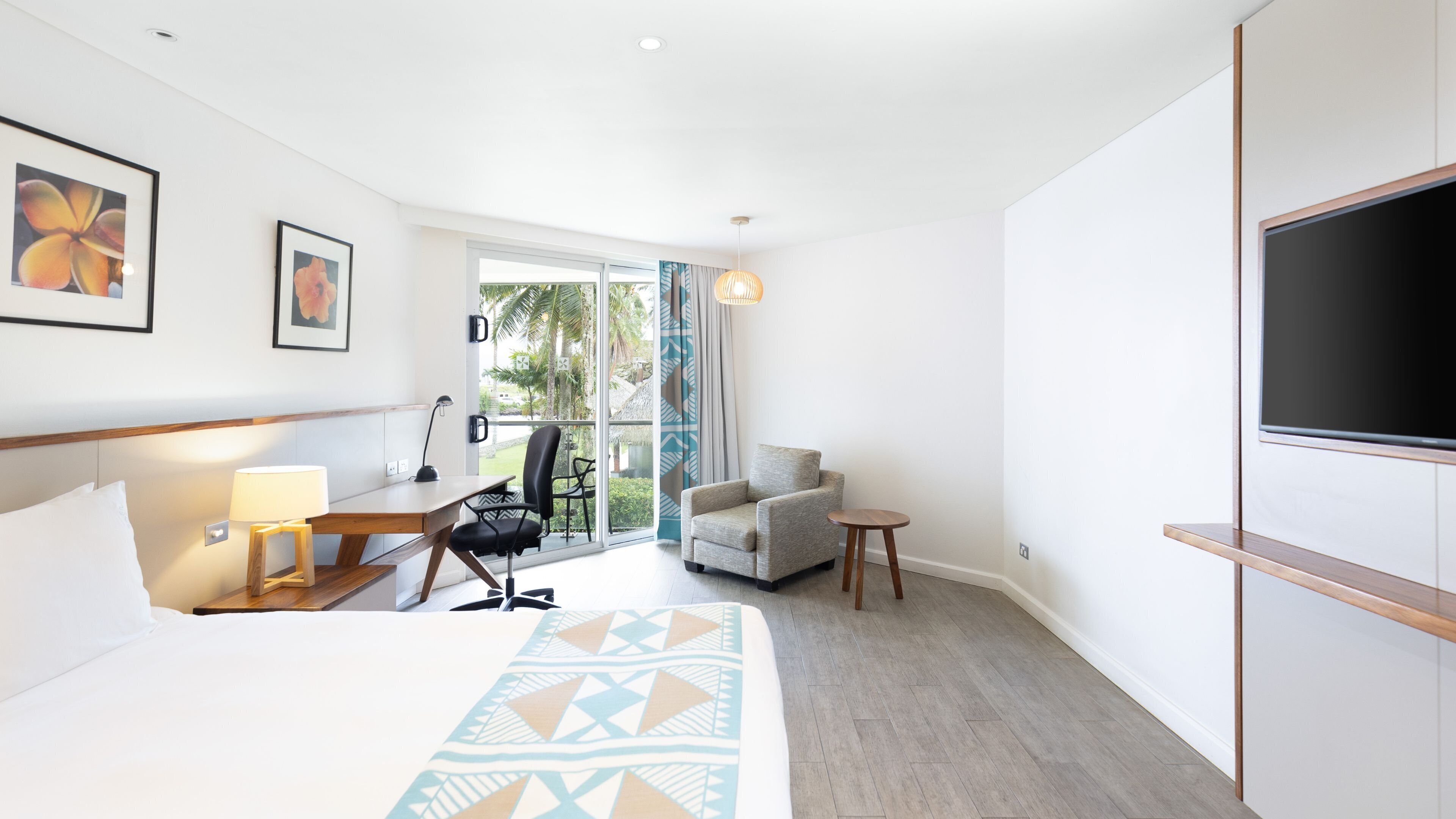 Habitación de hotel con cama, escritorio, silla, televisor y balcón con vistas a palmeras.