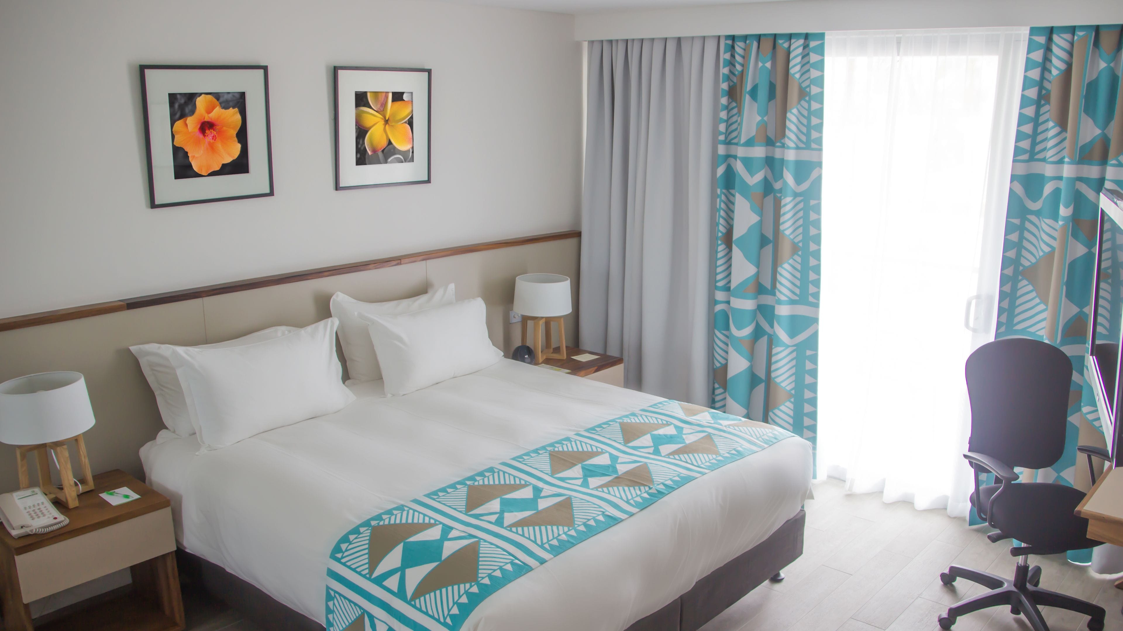 A hotel room with a large bed, two bedside tables with lamps, a desk with a chair, and a window with curtains.