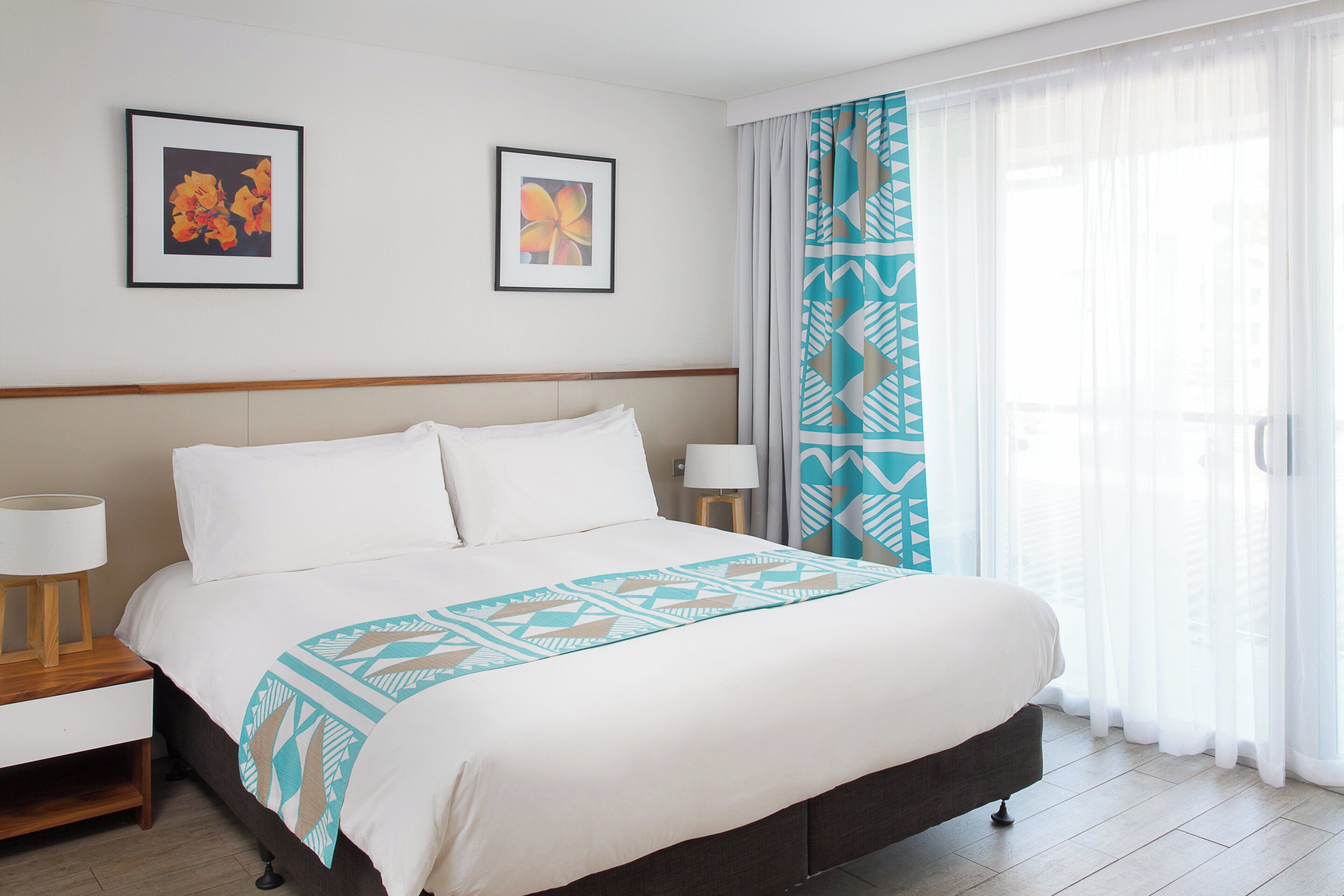 A hotel room with a large bed, two bedside tables, a nightstand, and a window with sheer curtains.