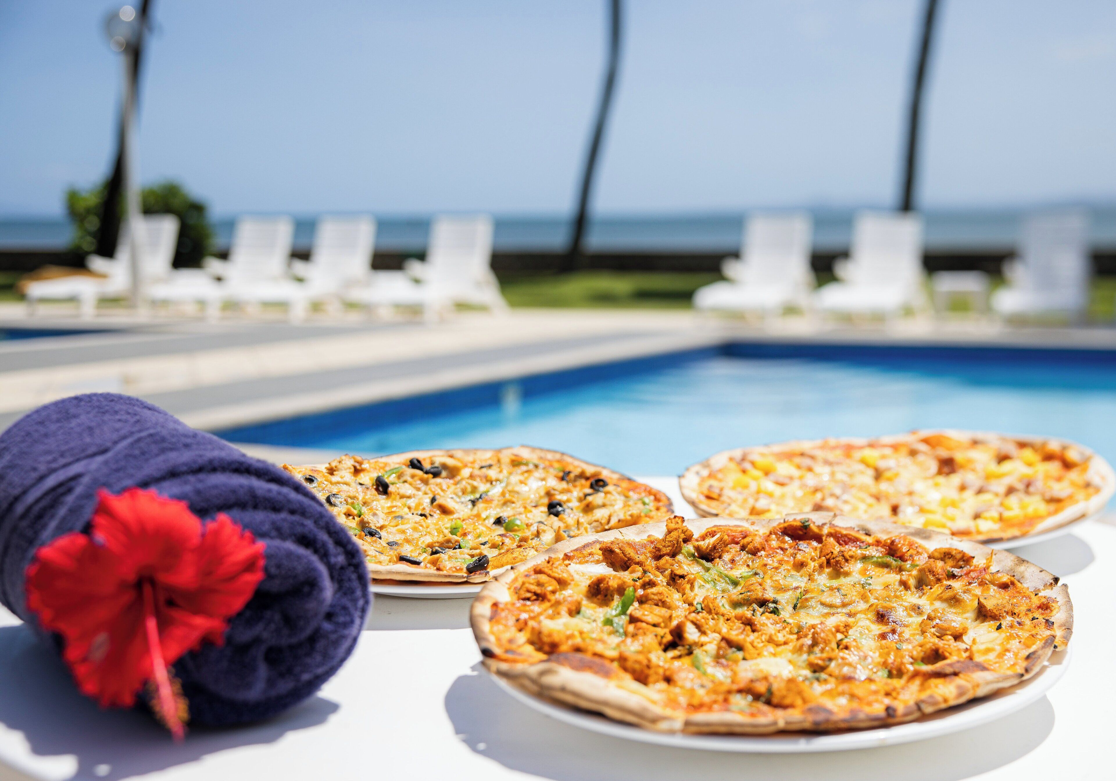 Three pizzas on a table near a pool with lounge chairs in the background.