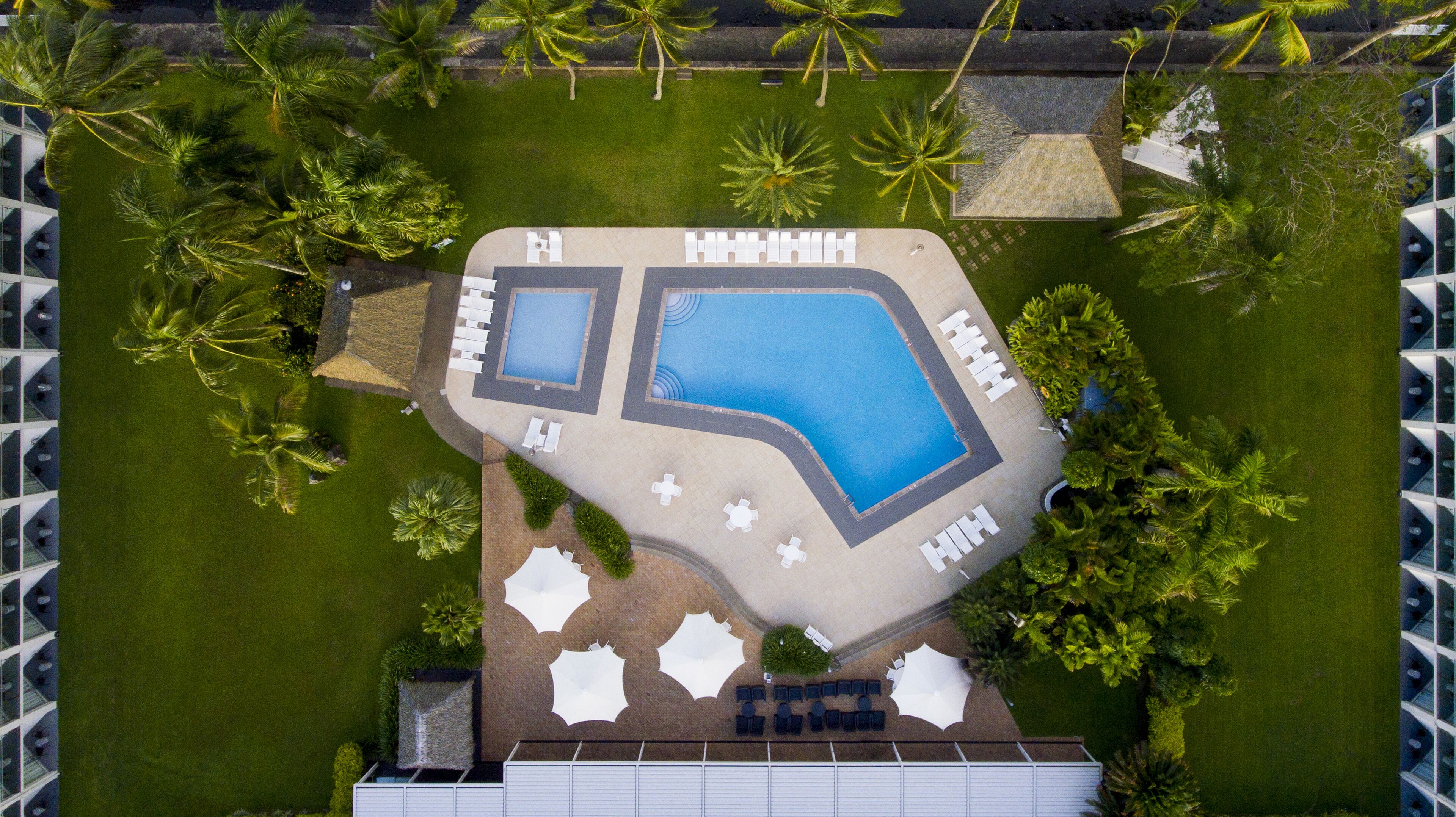 An aerial view of a resort with two swimming pools, sun loungers, and palm trees.