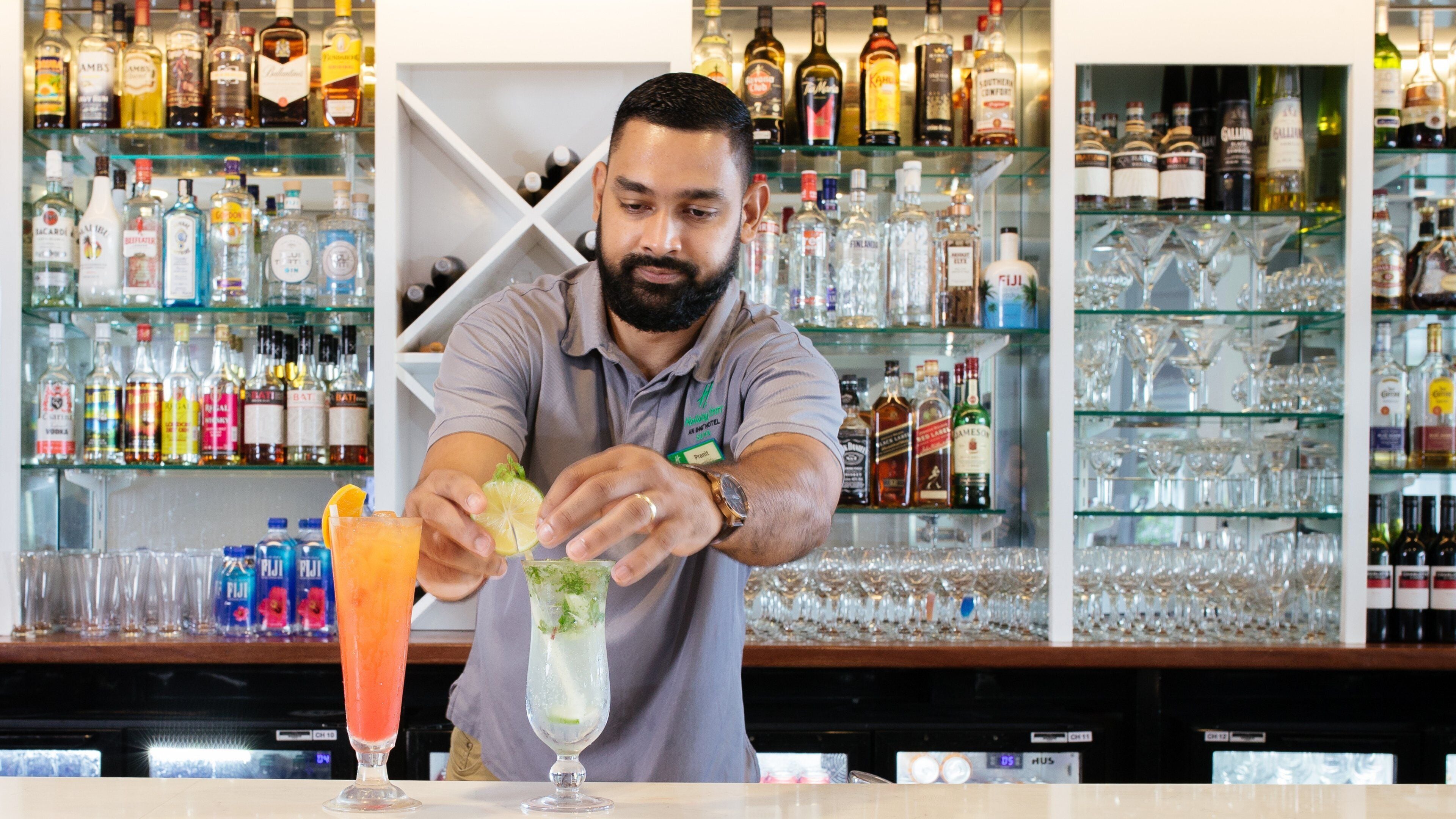 A bartender preparing a drink at a bar with shelves of liquor bottles and glasses in the background.
