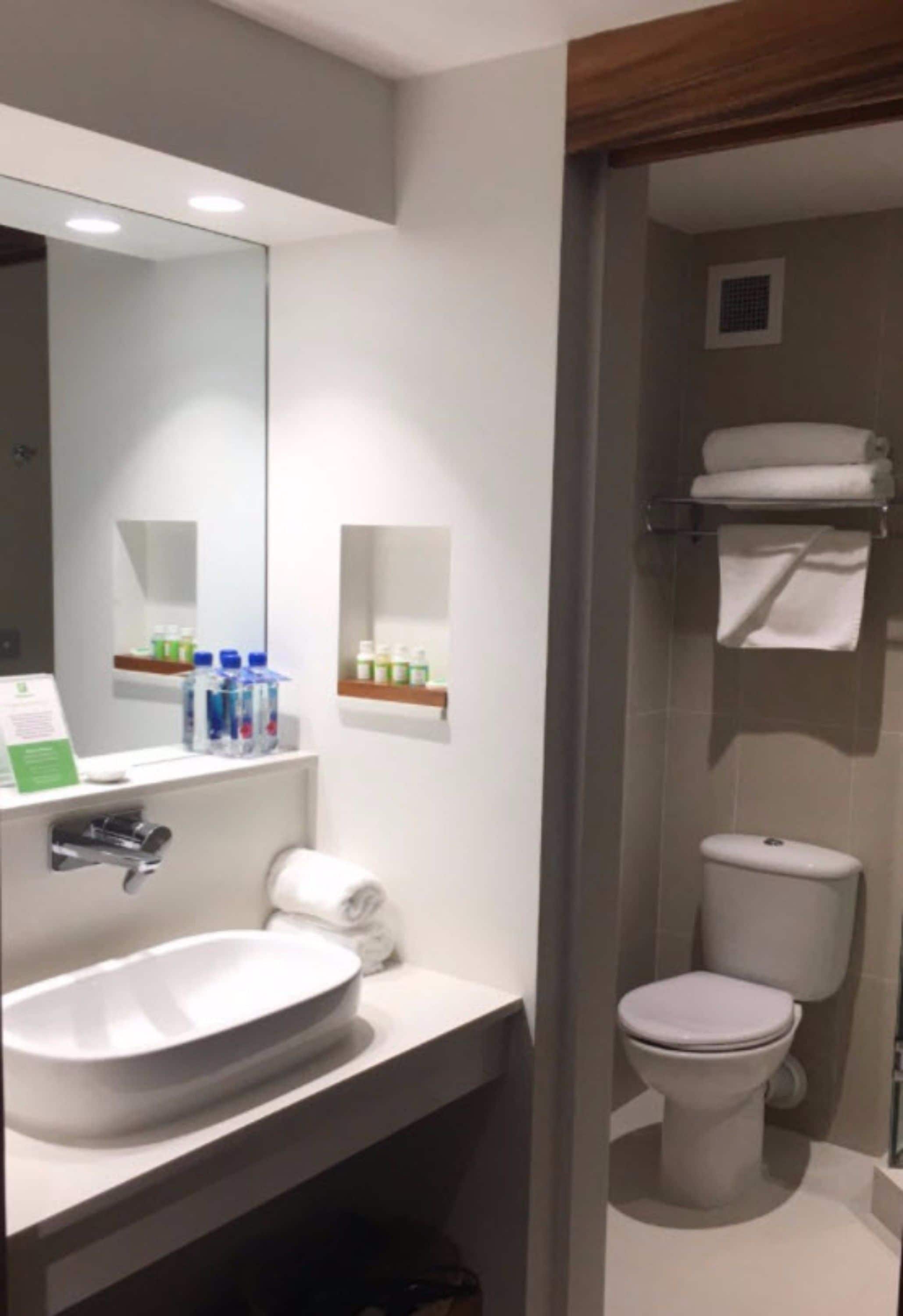 Une salle de bain moderne avec un lavabo blanc, un miroir et des toilettes. On y trouve des étagères avec des articles de toilette et un porte-serviettes au-dessus des toilettes.