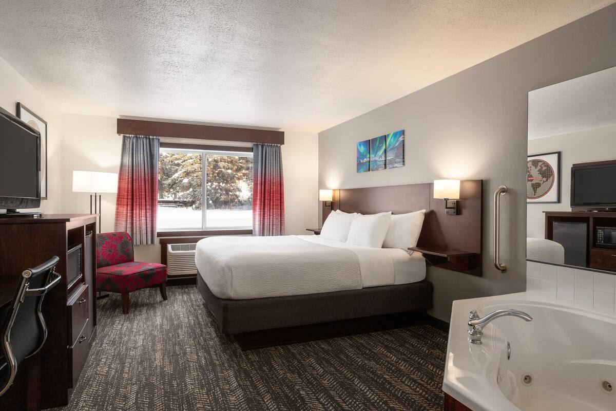 Een hotelkamer met een bed, een bureau met een televisie, een stoel, een raam met gordijnen, een tv aan de muur, een badkamer met bad en kunstwerken aan de muren.
