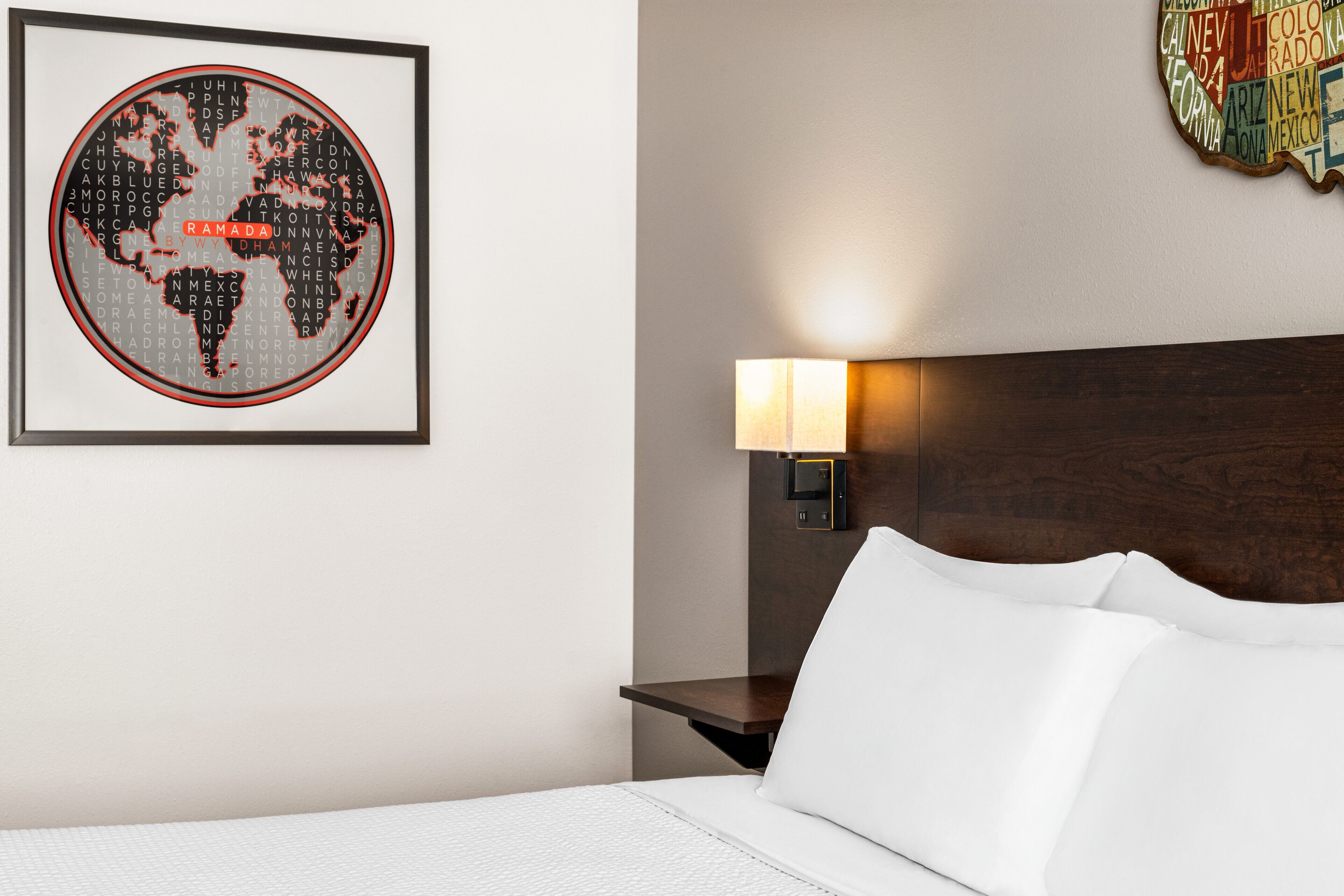 A hotel room with a neatly made bed, a framed world map artwork, a bedside lamp, and a wooden headboard.