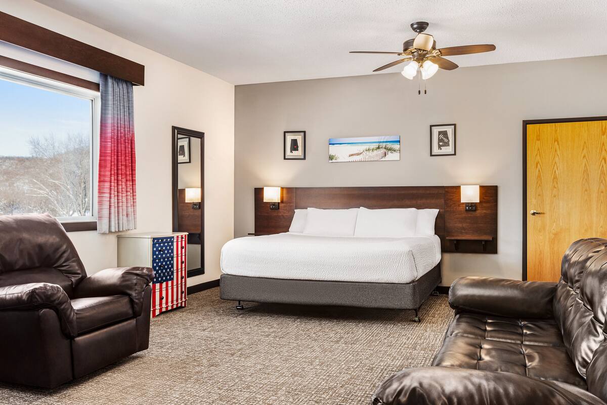 Een hotelkamer met een bed, twee leren banken, een plafondventilator en een groot raam met gordijnen.