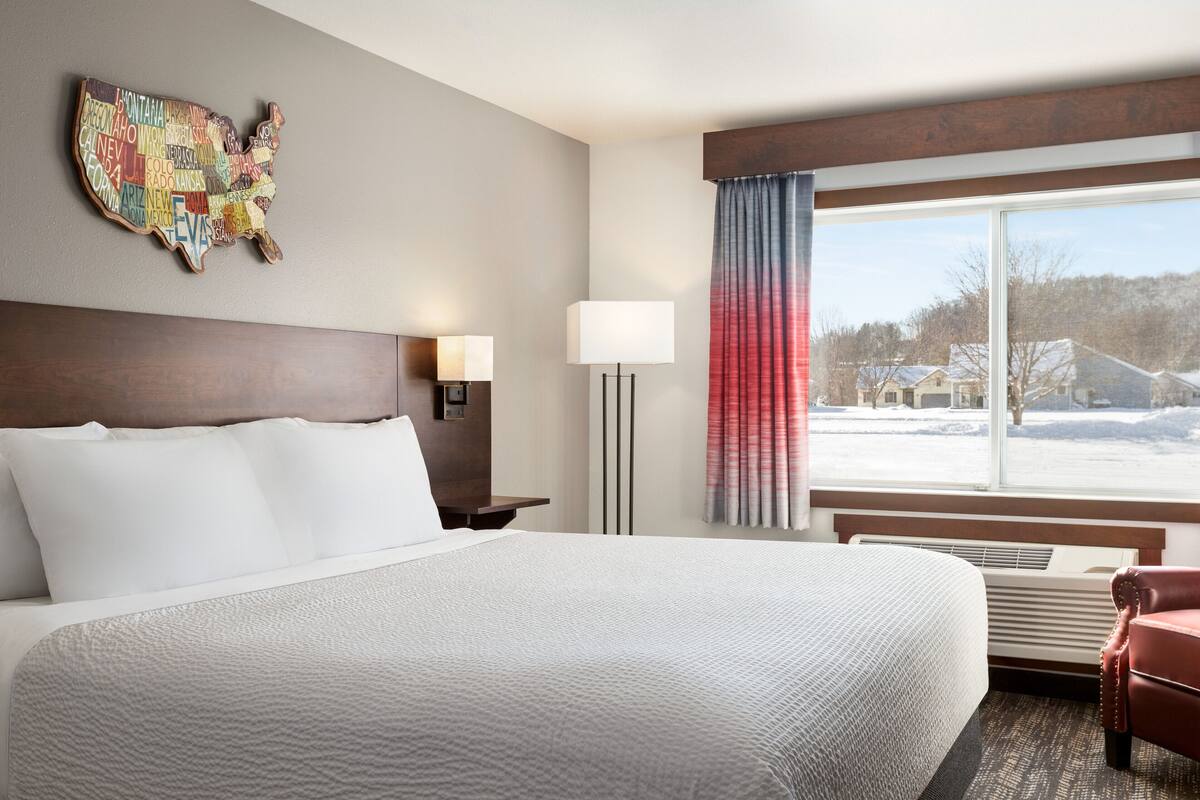 Een hotelkamer met een groot bed, een hoofdeinde, een nachtkastje, een lamp, een raam met uitzicht op huizen en een wereldkaart.