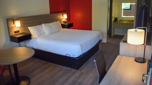 Habitación de hotel con cama, mesitas de noche, escritorio, silla, lámpara y baño a la vista a través de una puerta abierta.