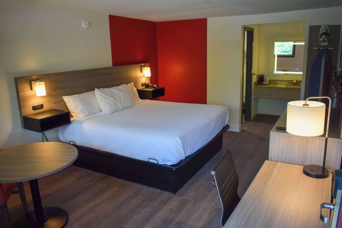 Habitación de hotel con cama, mesitas de noche, escritorio, silla, lámpara y baño a la vista a través de una puerta abierta.