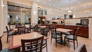 Un comedor amplio con varias mesas y sillas, una cocina al fondo y un ventanal que proporciona luz natural.