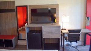 Una habitación de hotel moderna con escritorio, televisión, refrigerador pequeño, silla y mesita de noche.