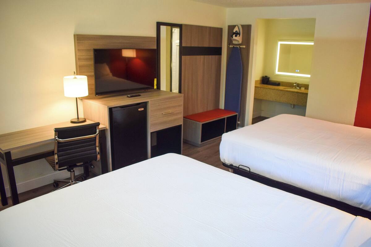 Habitación de hotel con dos camas, un escritorio con lámpara, televisor, un pequeño espacio de descanso y baño con espejo y lavabo.