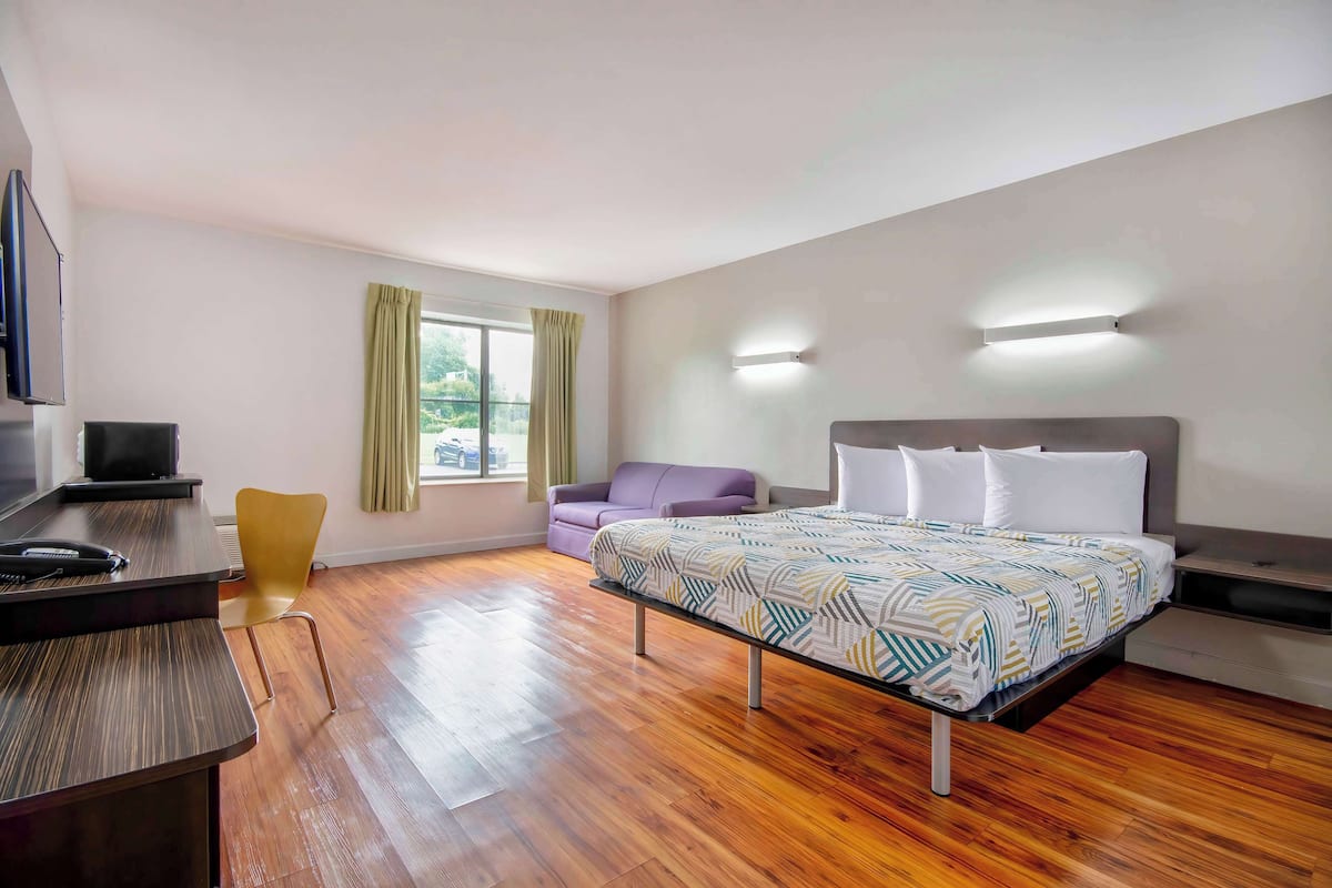 A modern hotel room with a wooden floor, a bed with patterned bedding, a desk with a chair, a sofa, and a television.