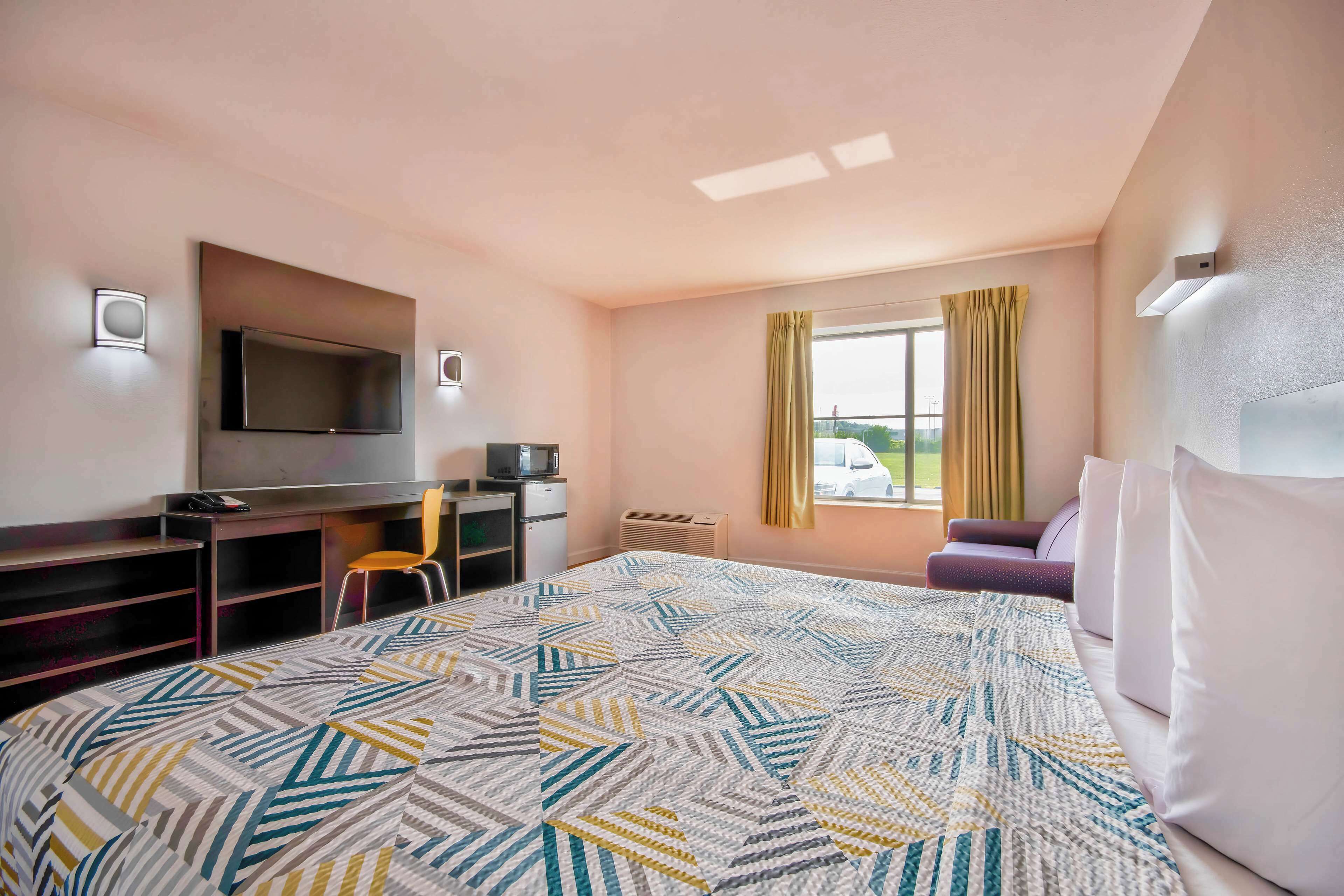 A hotel room with a bed, a desk with a TV, a chair, a window with curtains, and a view of the outside.