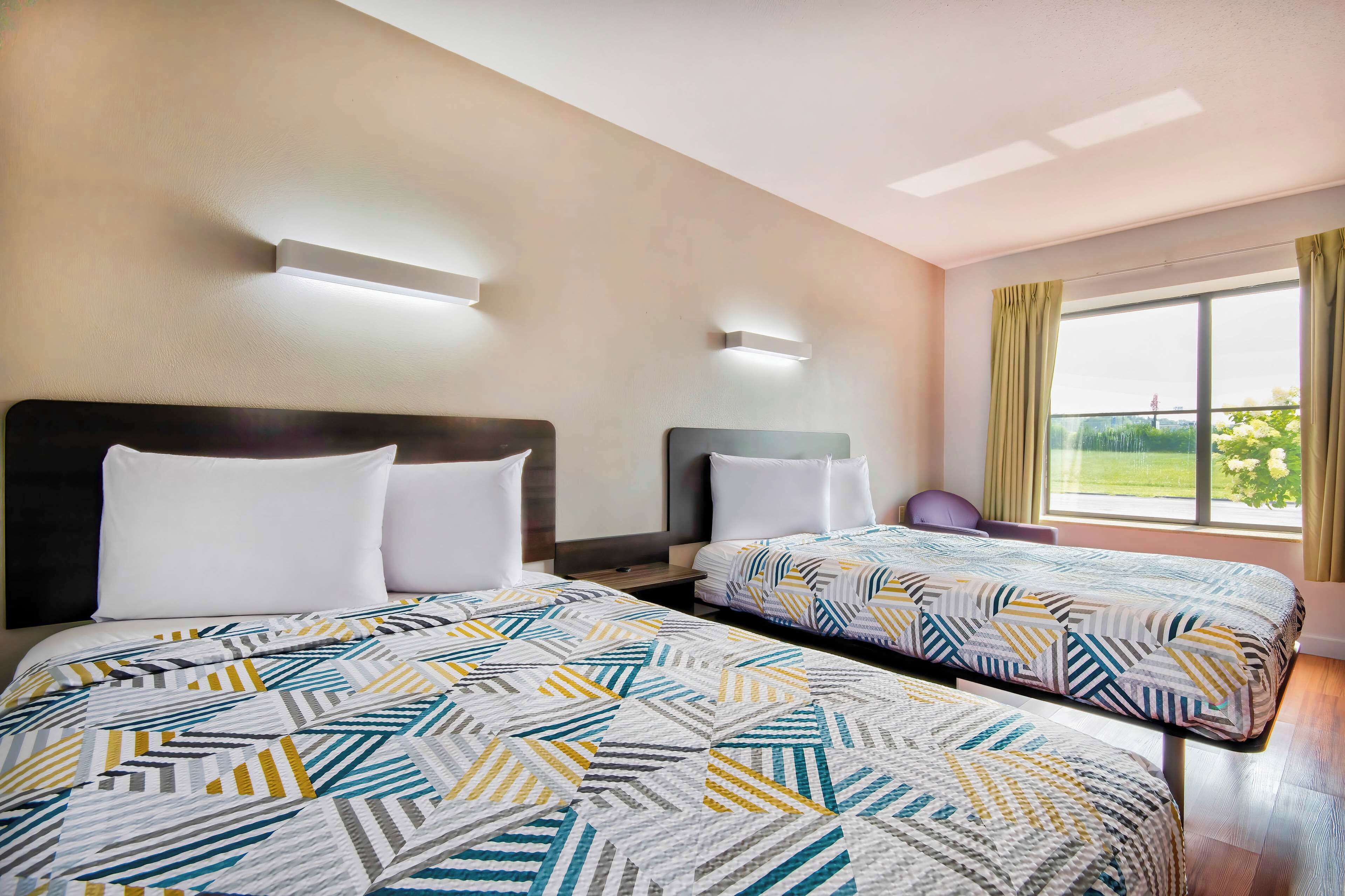 Une chambre d’hôtel avec deux lits, chacun recouvert d’une couvre-lit à motifs, une table de chevet, une lampe fixée au mur et une grande fenêtre avec des rideaux.