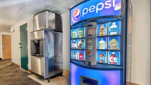 Máquina expendedora con logotipo de Pepsi y varias opciones de bebidas, un refrigerador de acero inoxidable y una puerta al fondo.