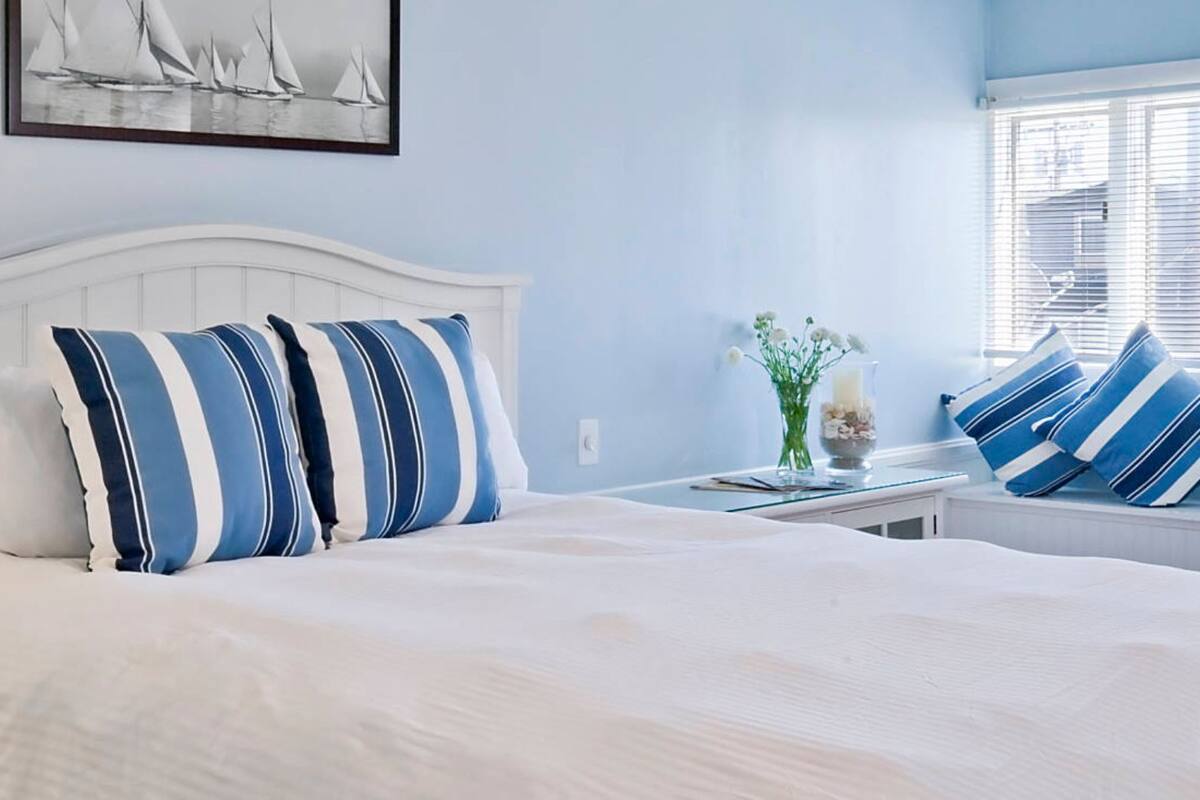 Een netjes opgemaakt bed met witte beddengoed en blauw gestreepte kussens, een bedlampje, een telefoon en een ingelijste foto aan de muur.